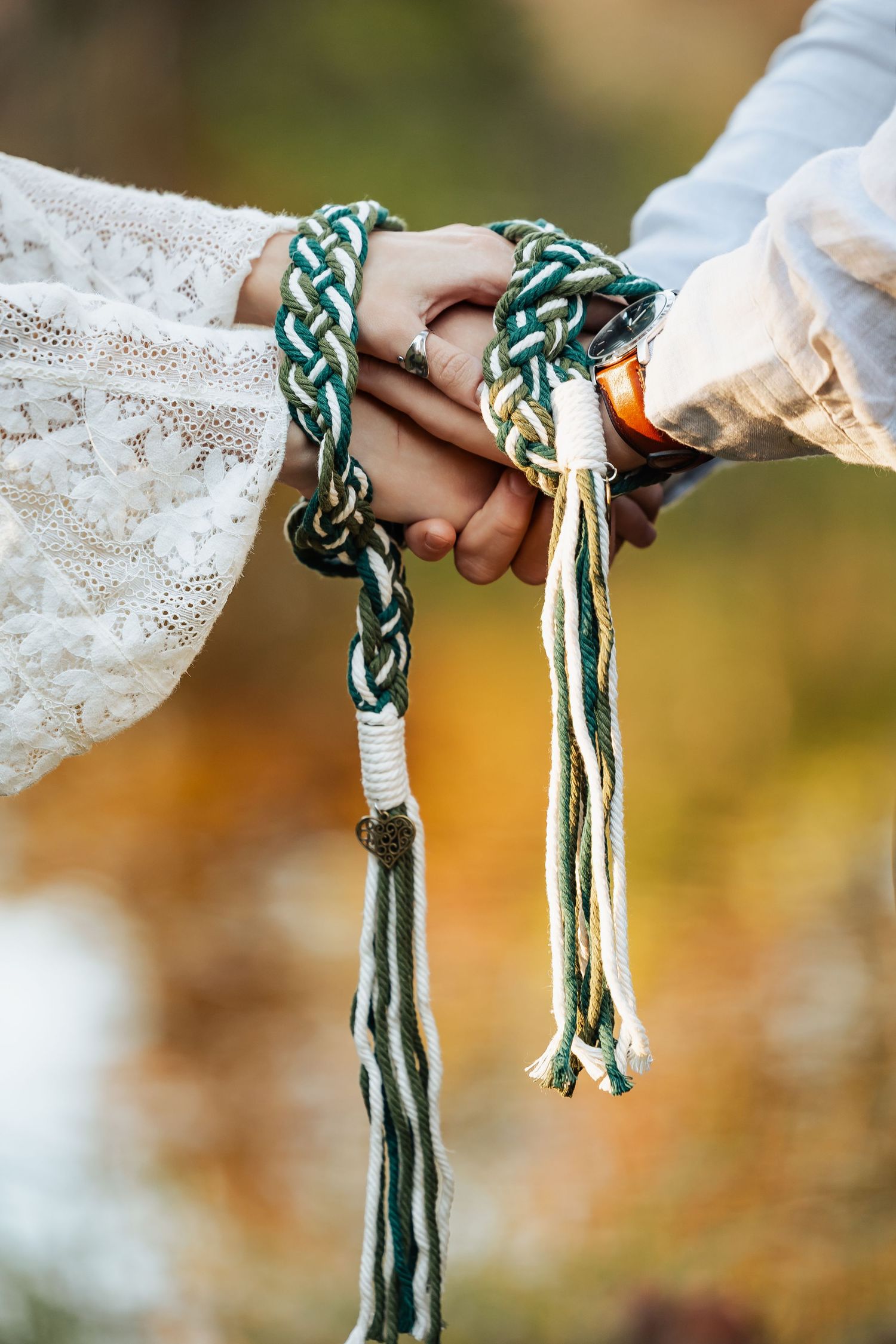Close-up of two joined hands wrapped in a green and white braided handfasting cord outdoors.