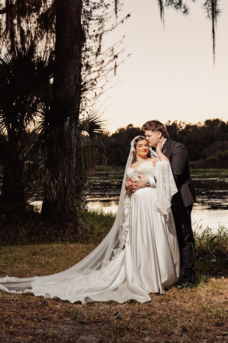 Groom kissing bride on cheek as she smiles in lace wedding gown by a lakeside at sunset.