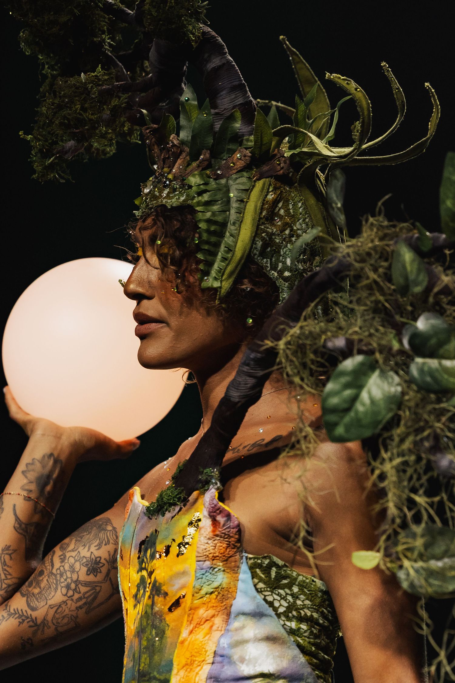 Close-up of tattooed model in nature-themed costume holding glowing sphere.