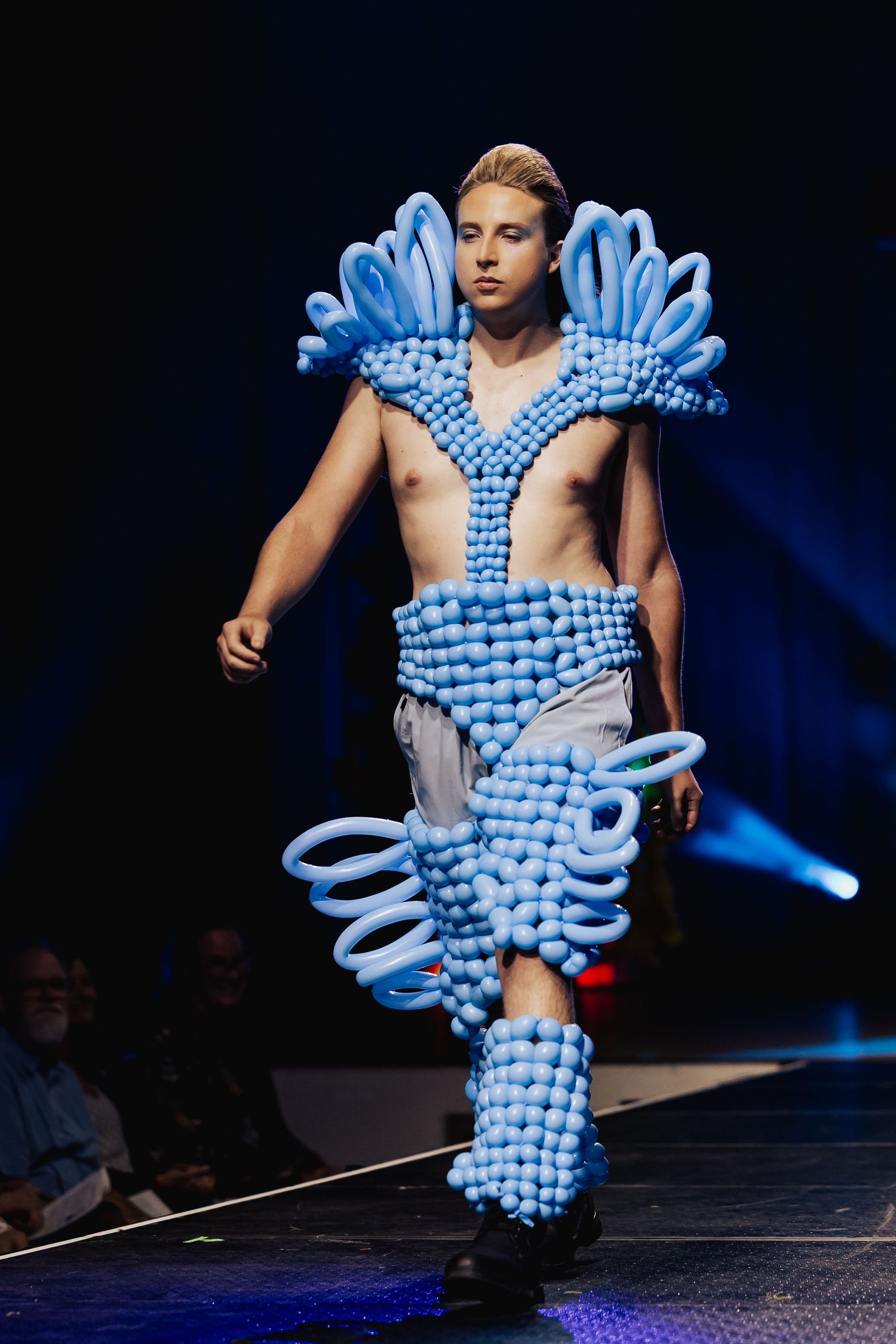 Male model walks runway wearing blue balloon art ensemble with sculptural collar and leg details.