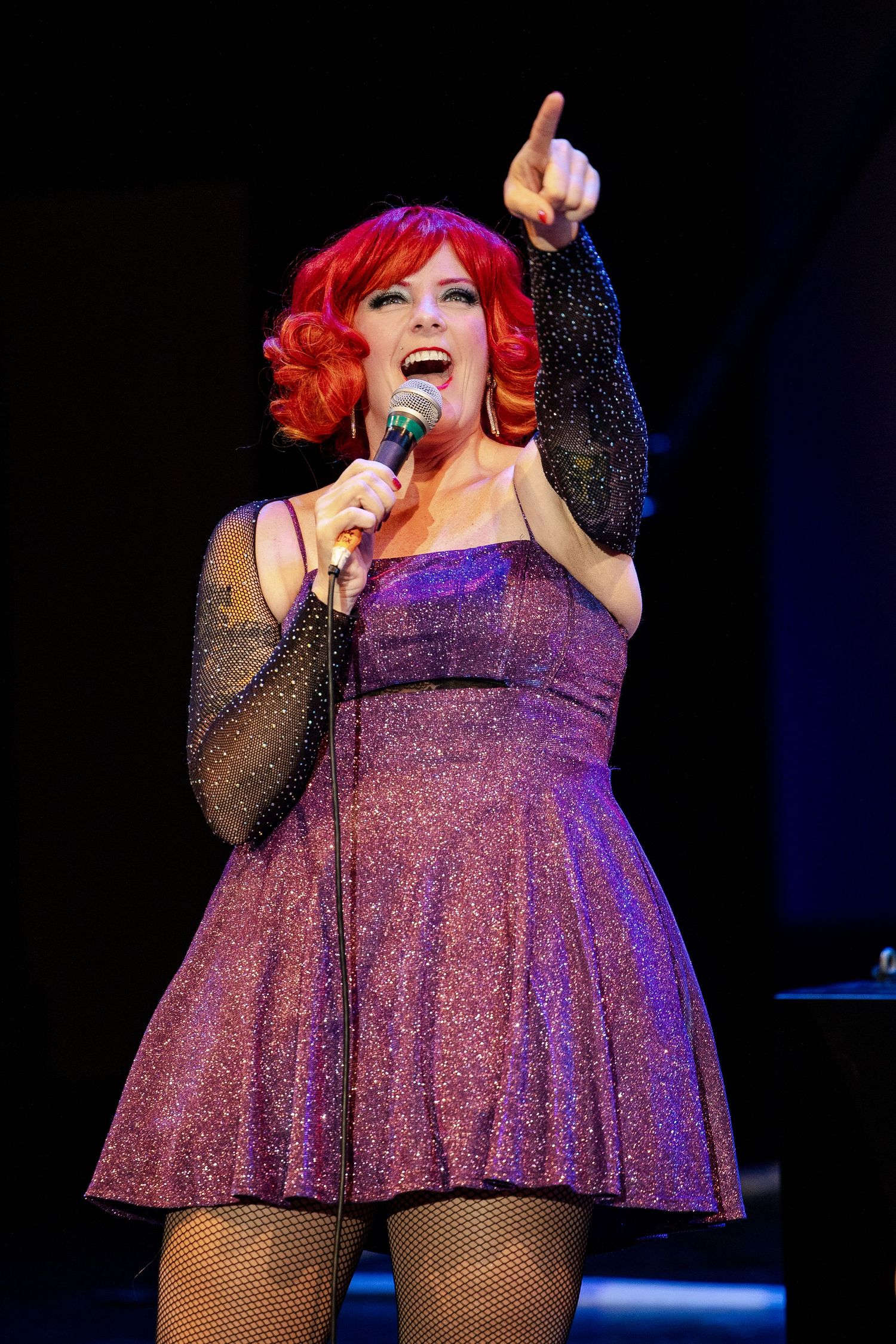 Red-haired female performer in purple sparkle dress singing and pointing upward on dark stage.