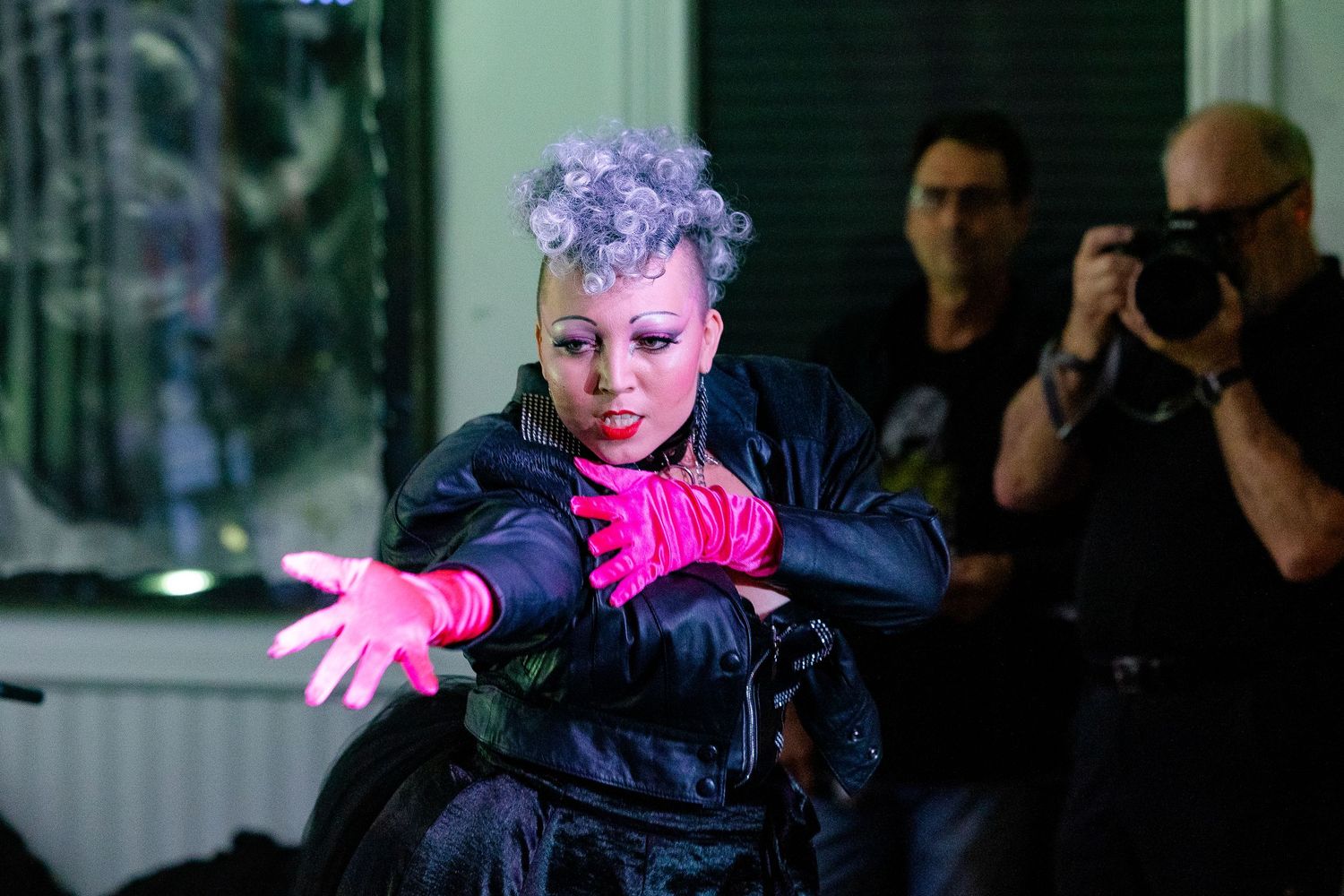 Performer with purple hair and pink gloves in black jacket dancing on stage with photographers in background.