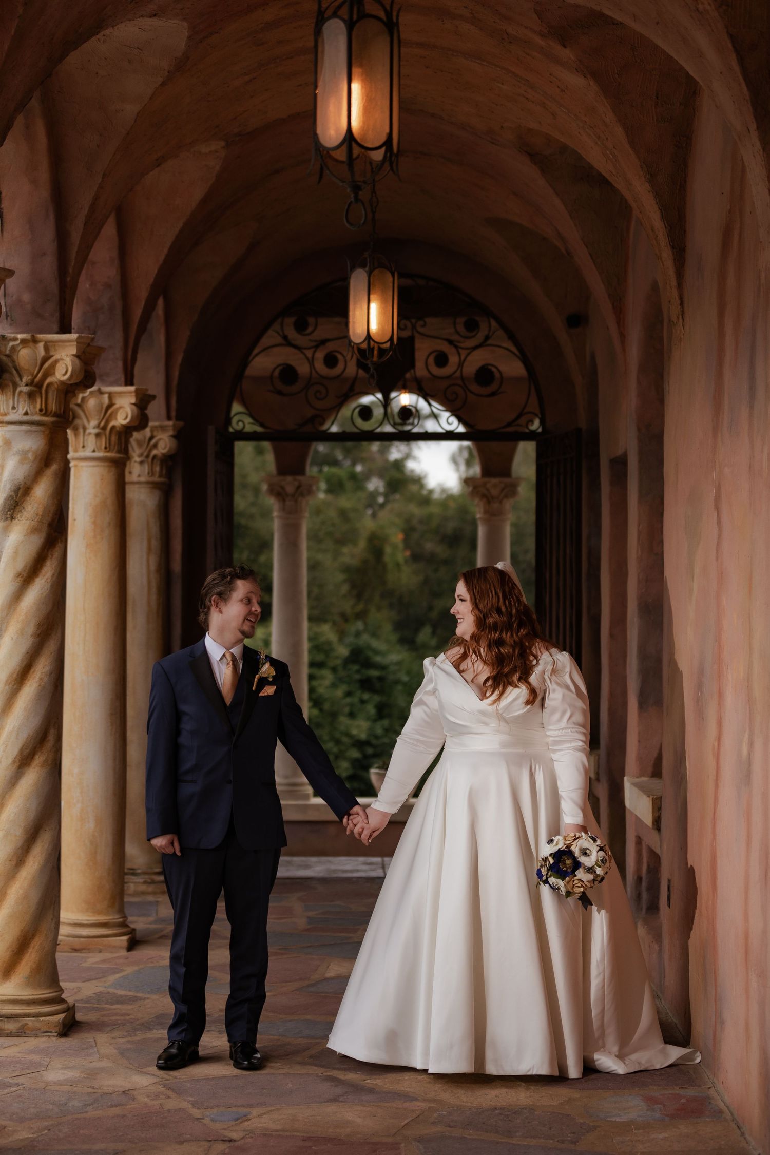 Bride and groom hold hands under ornate archway with hanging lantern, bride in white gown holding bouquet.