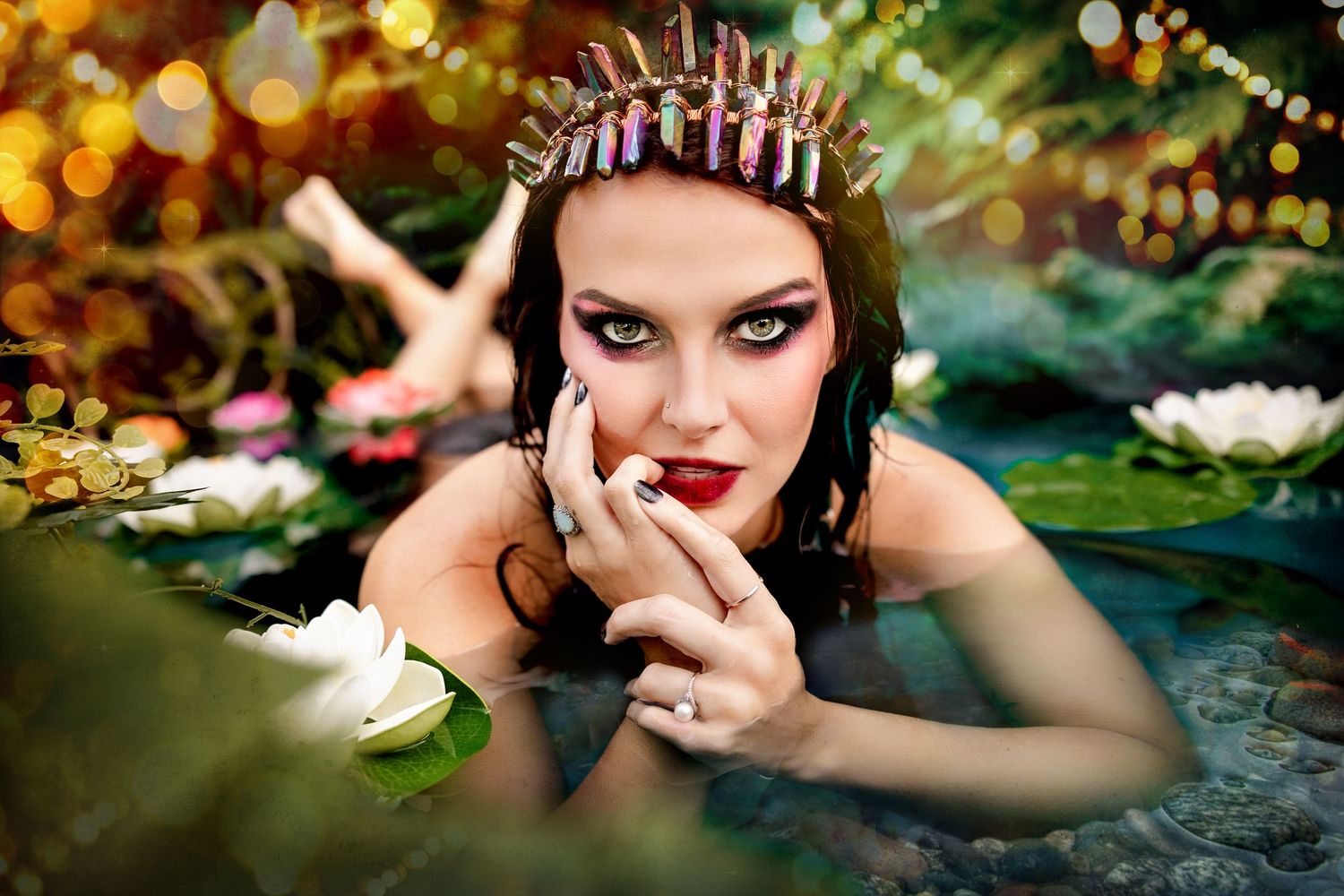 Close-up of woman with dramatic makeup and crystal crown posing in water with white lotus flowers.