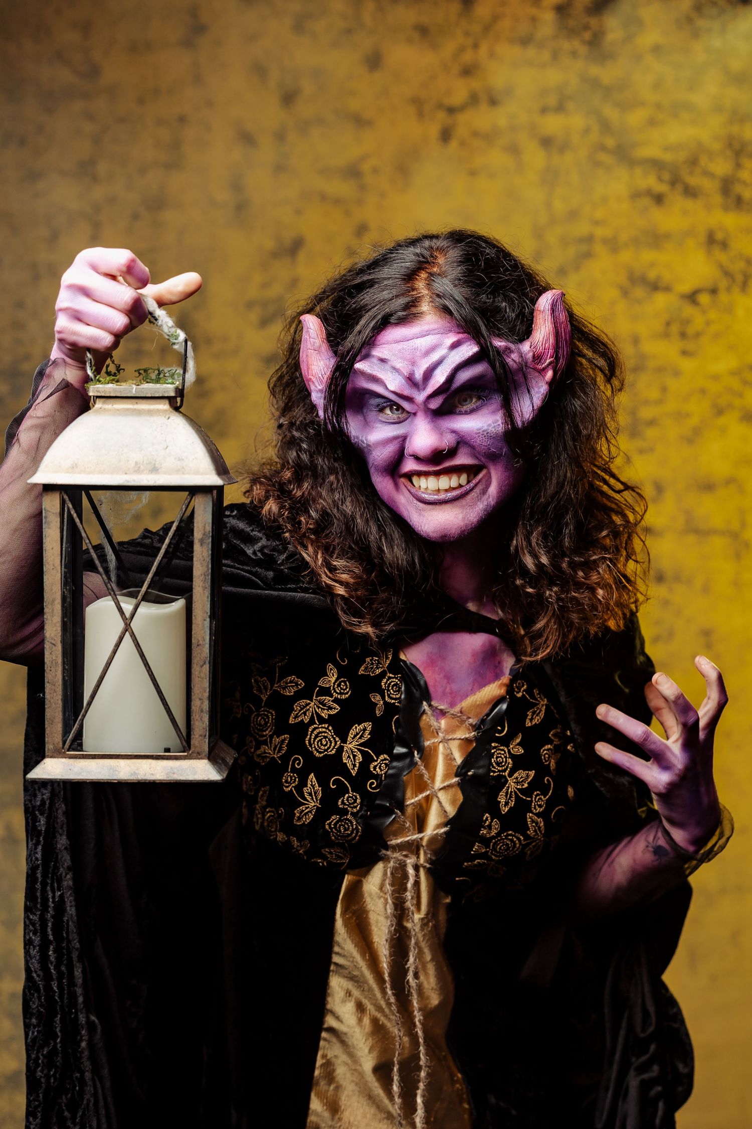 Person in purple demon Halloween makeup with horns holds a candle lantern against a yellow background.