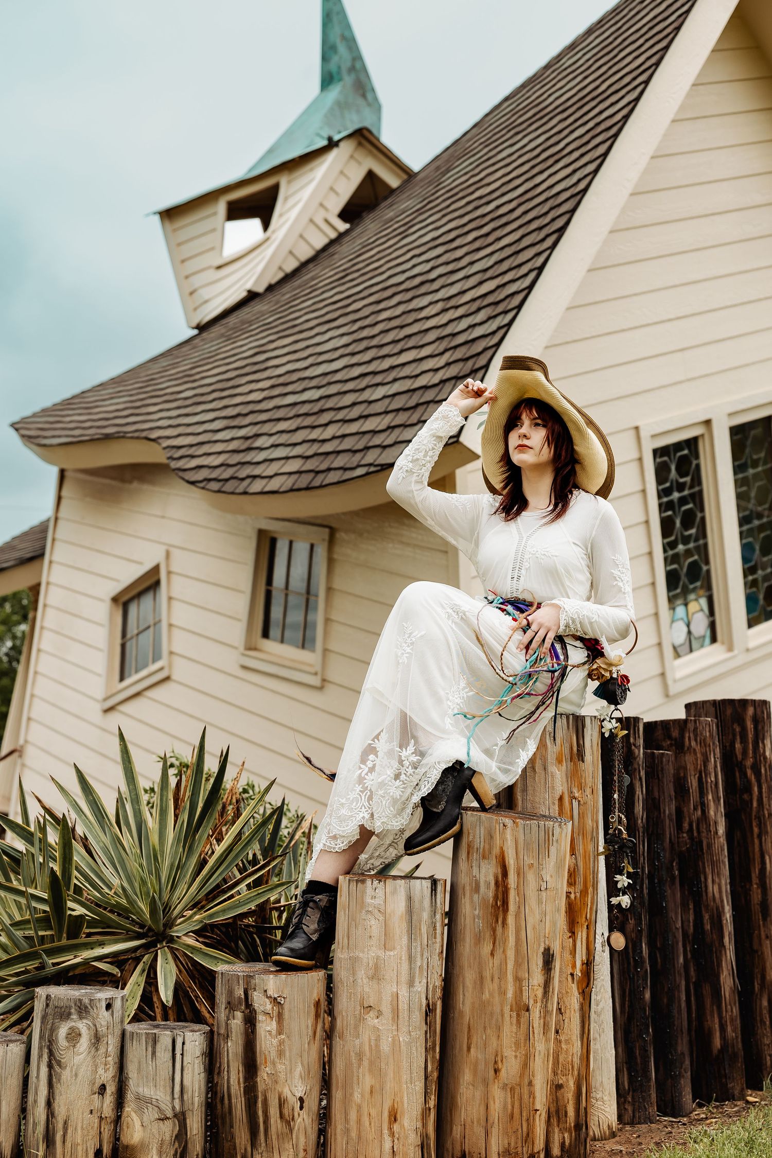 Woman in white lace dress and straw hat sits atop wooden fence posts before a unique chapel.