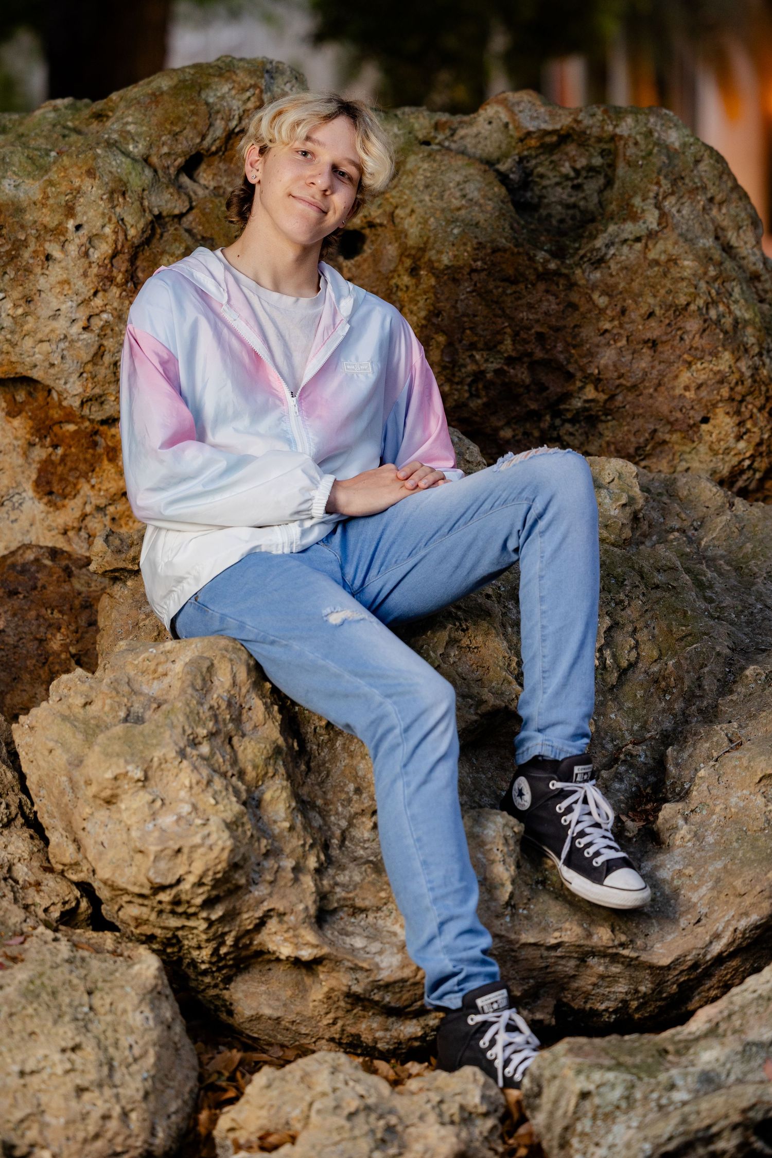 Teen with blonde hair sits on rocks wearing tie-dye hoodie, blue jeans, and black Converse sneakers outdoors.