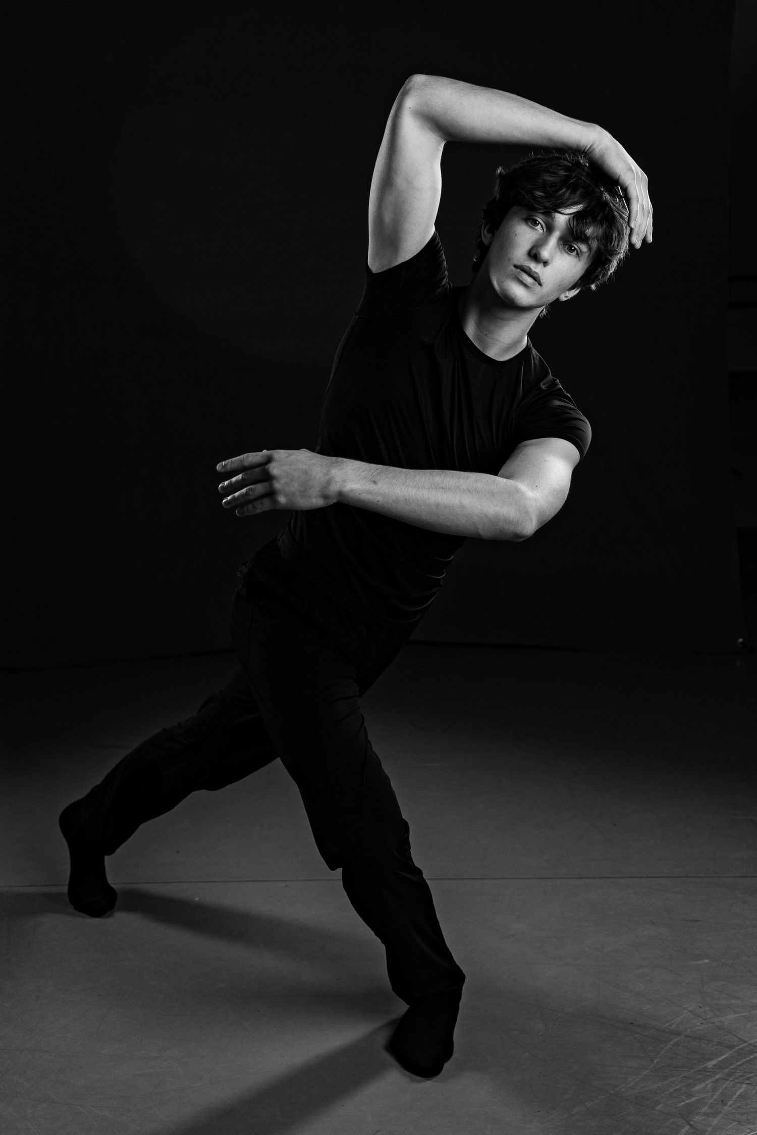 Male dancer in black outfit strikes dramatic pose against dark background in black and white studio photo.