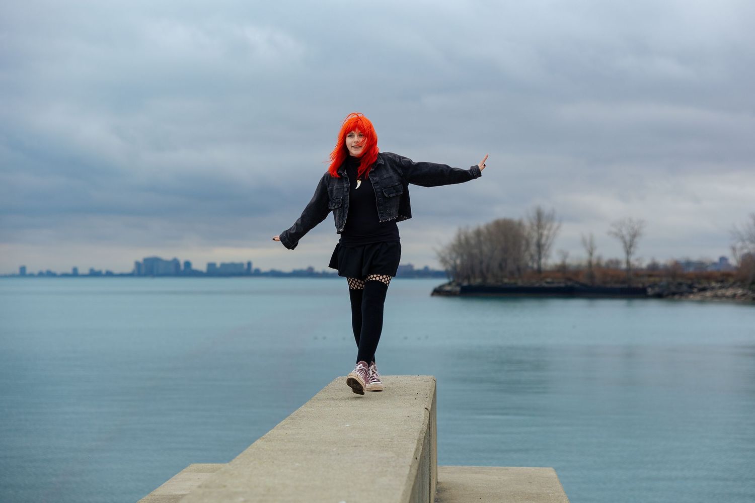 Woman with bright red hair balancing on concrete ledge by blue lake under cloudy sky wearing black jacket.