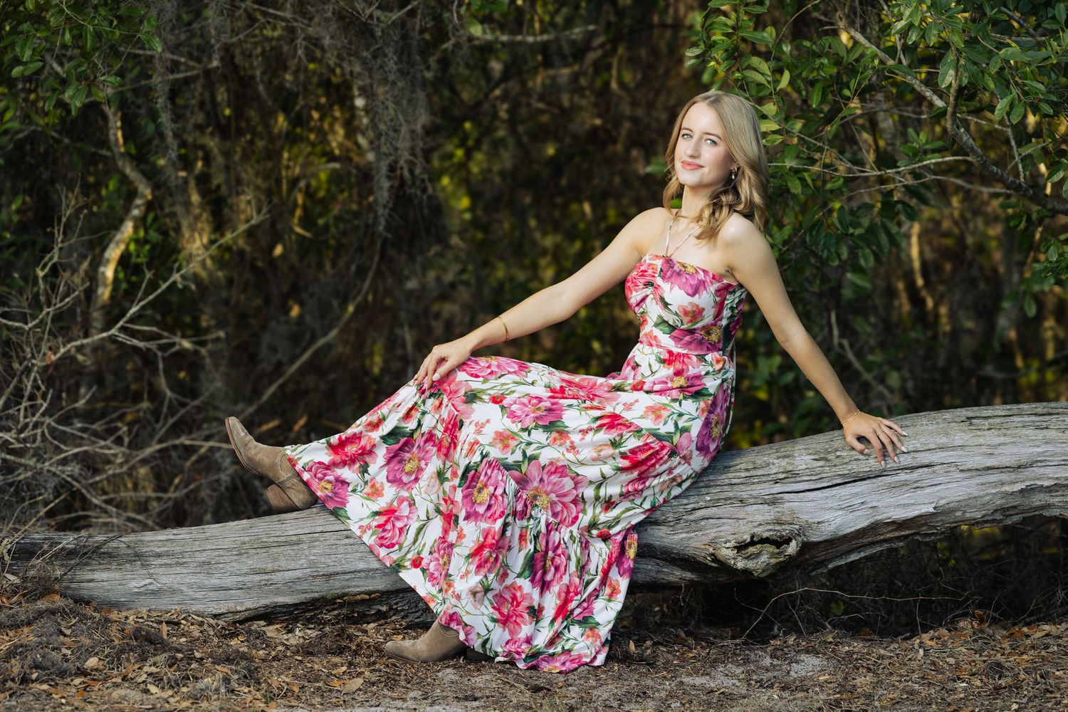 Young woman in floral maxi dress sitting gracefully on a fallen log in a lush forest setting.
