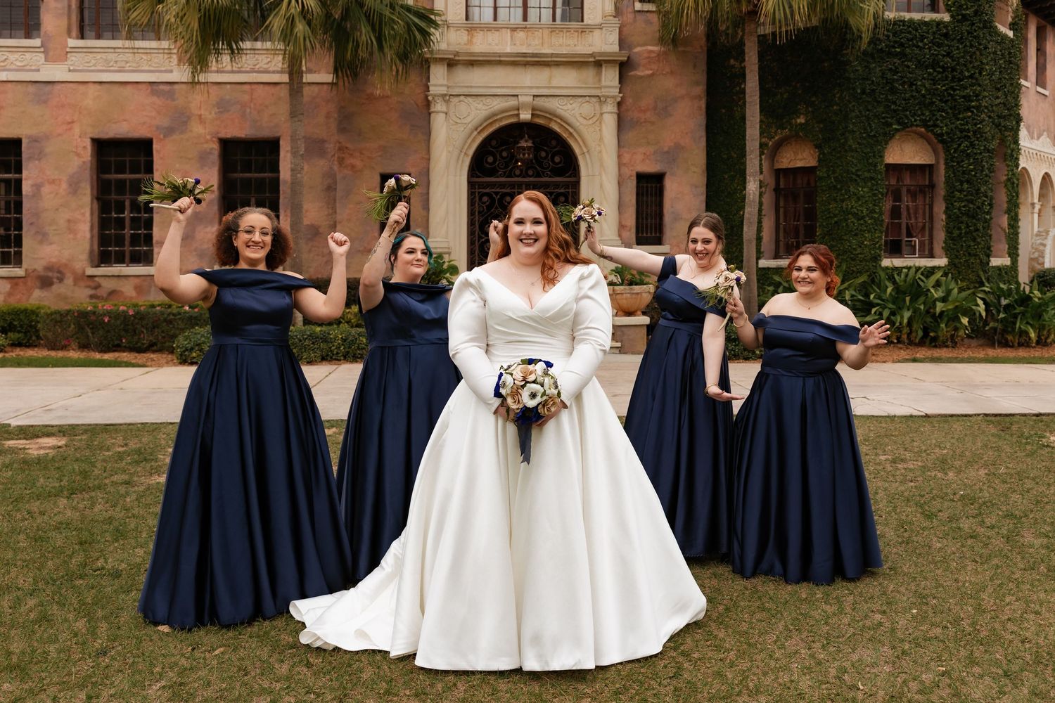 Bride in white gown celebrates with four bridesmaids in navy off-shoulder dresses outside an ivy-covered mansion.