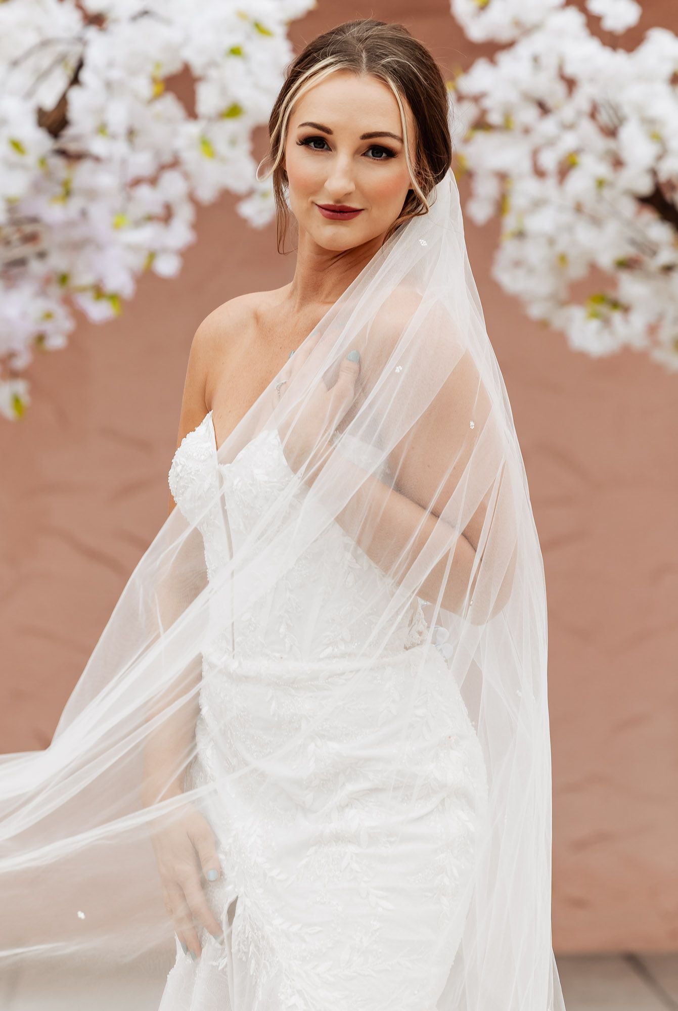Beautiful bridal portrait with flowing white wedding dress and veil against pink wall with cherry blossoms.