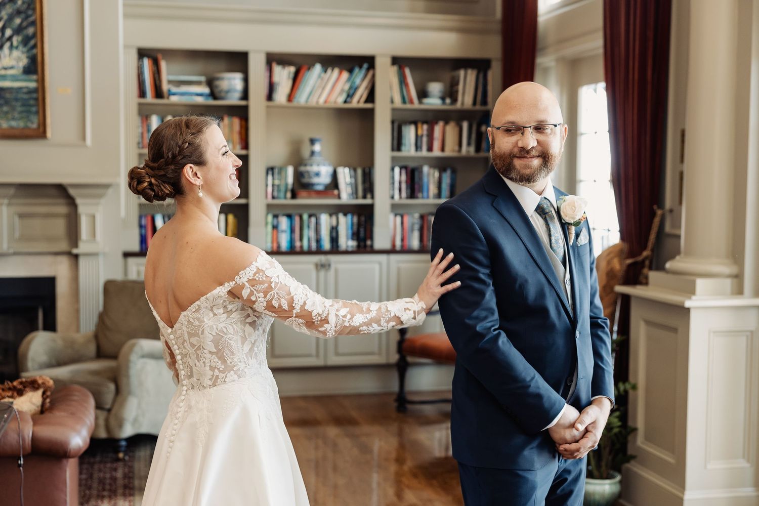 Bride in lace gown surprises groom in navy suit during wedding first look in classic library.