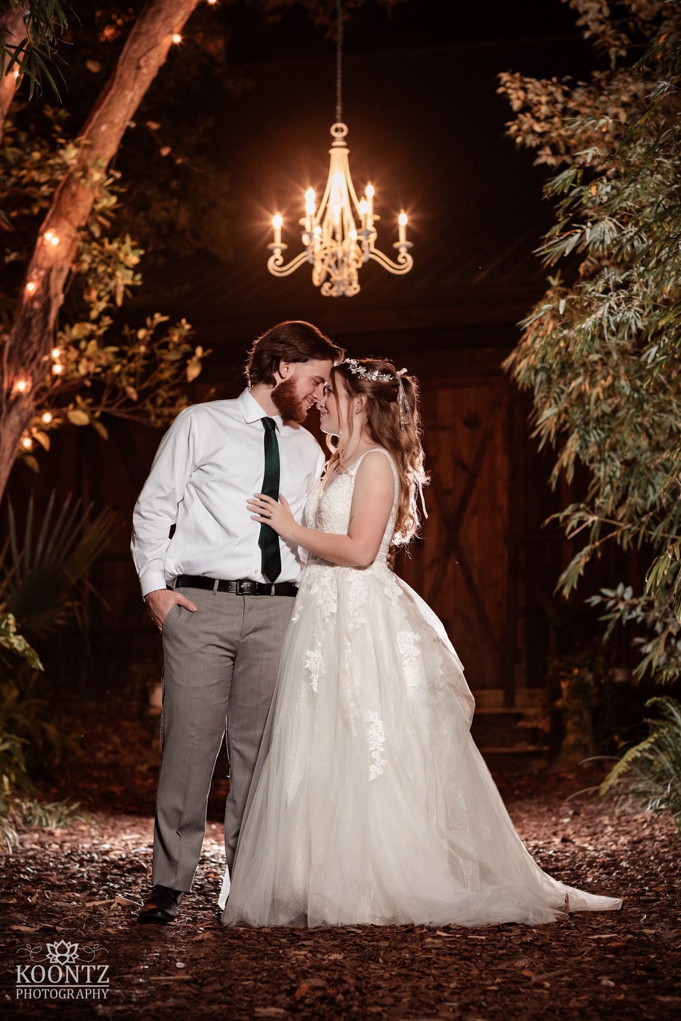 Newlyweds share a kiss under elegant chandelier surrounded by lush greenery at night wedding venue.
