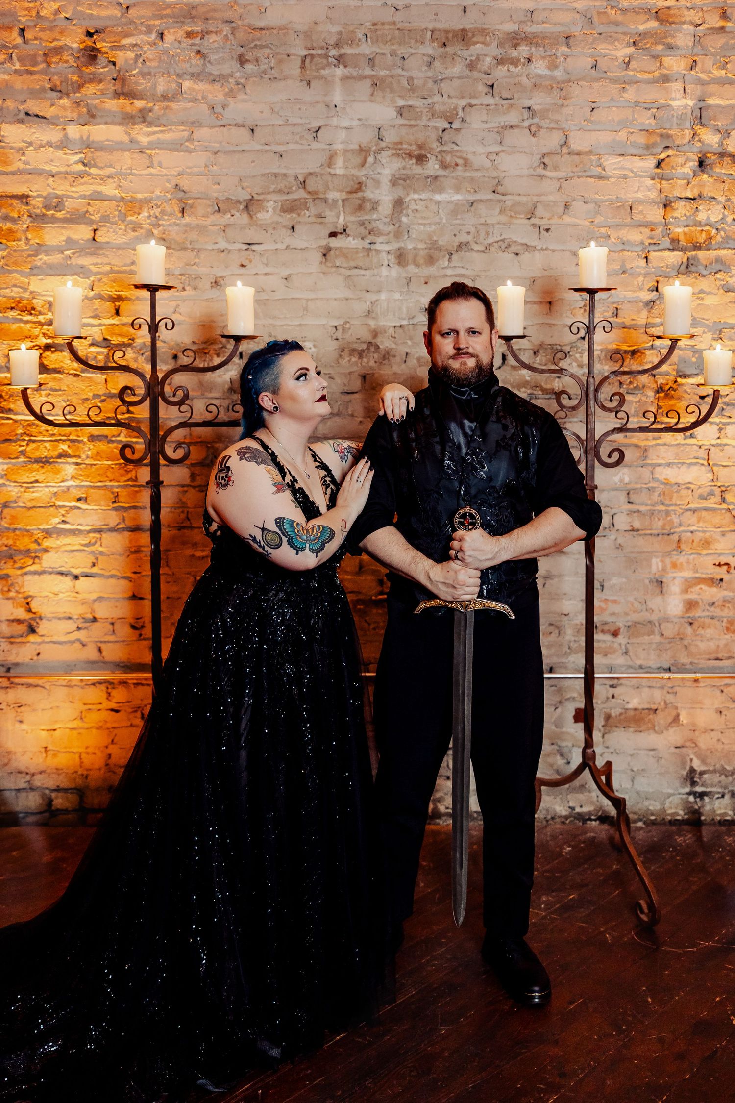 Gothic wedding couple posing with ornate sword before brick wall with candelabras and candles.
