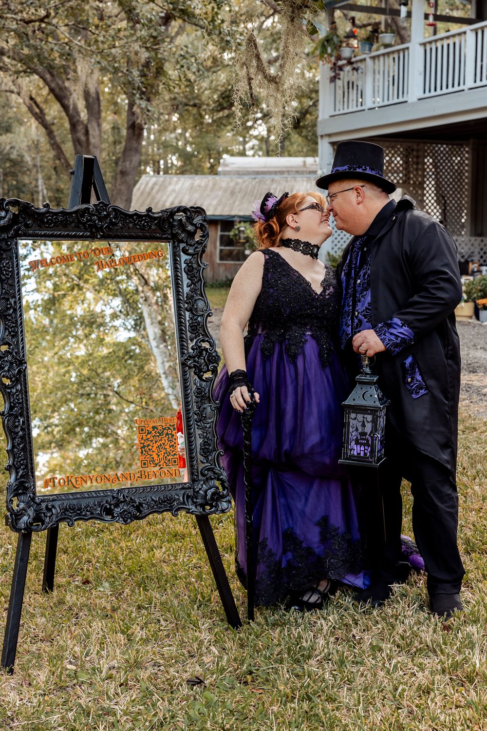 Gothic wedding couple in purple and black attire kiss beside ornate black mirror sign outdoors.