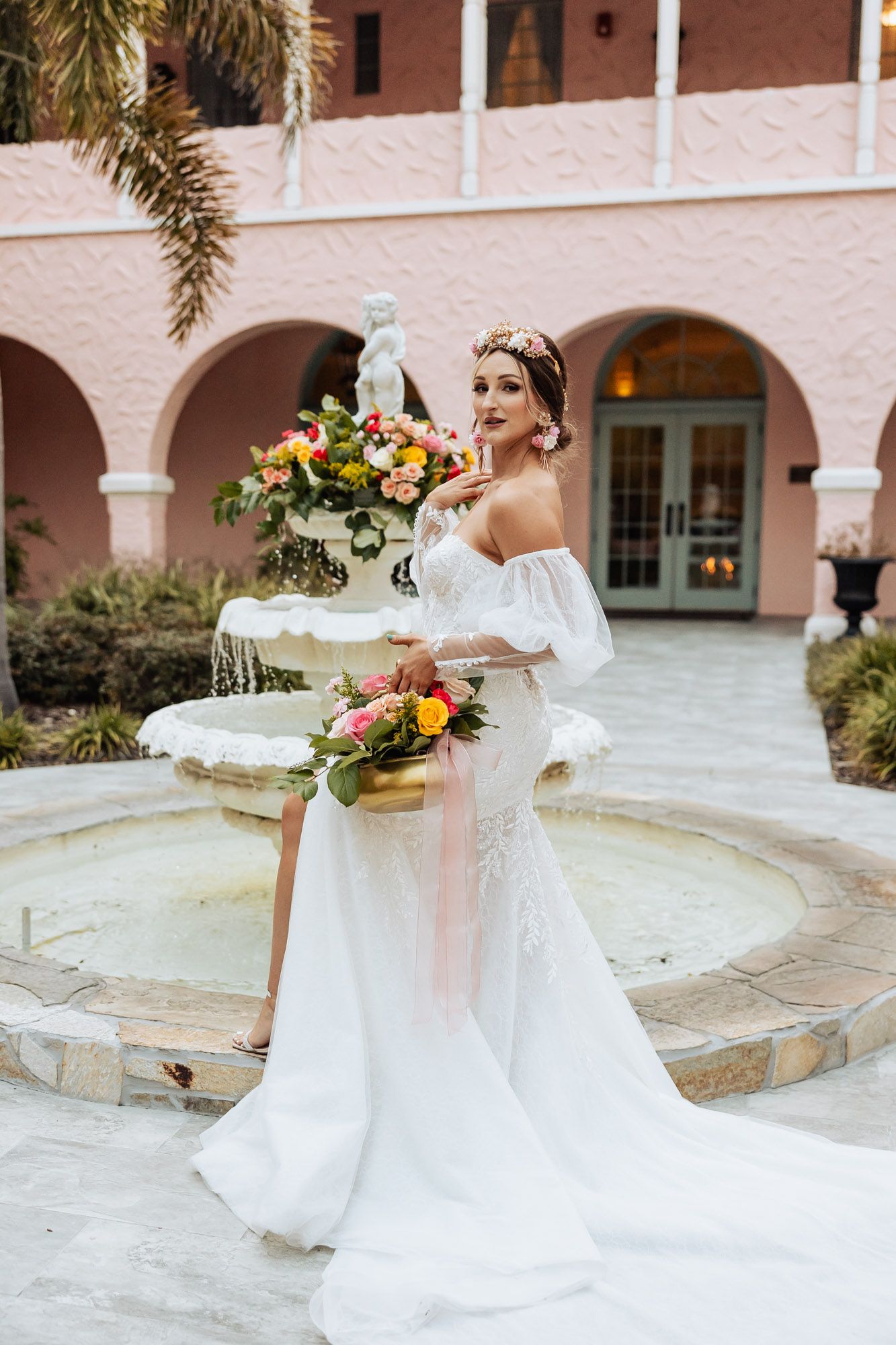 Elegant bride in off-shoulder white gown holds colorful bouquet beside ornate fountain at pink venue.