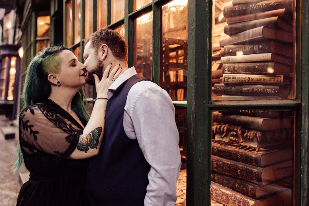 Couple sharing a kiss in front of a bookshop window filled with stacked books in Diagon Alley.