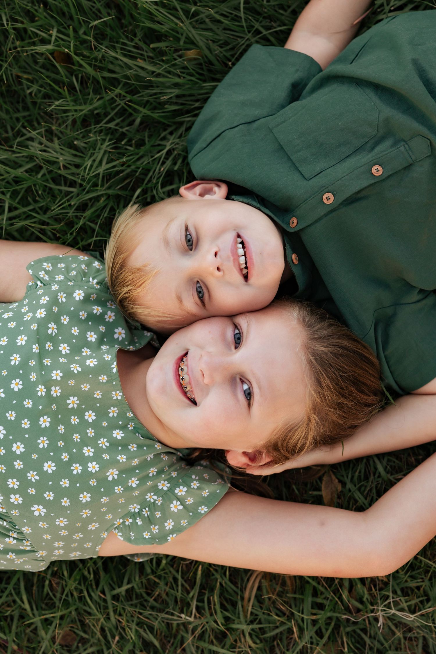 Two children lay head-to-head in grass wearing green and floral outfits.