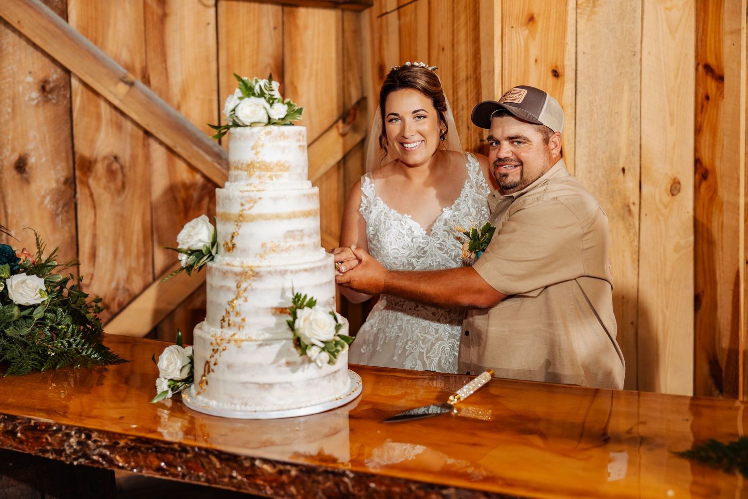 Wedding couple cuts elegant white tiered cake adorned with flowers in rustic barn venue.