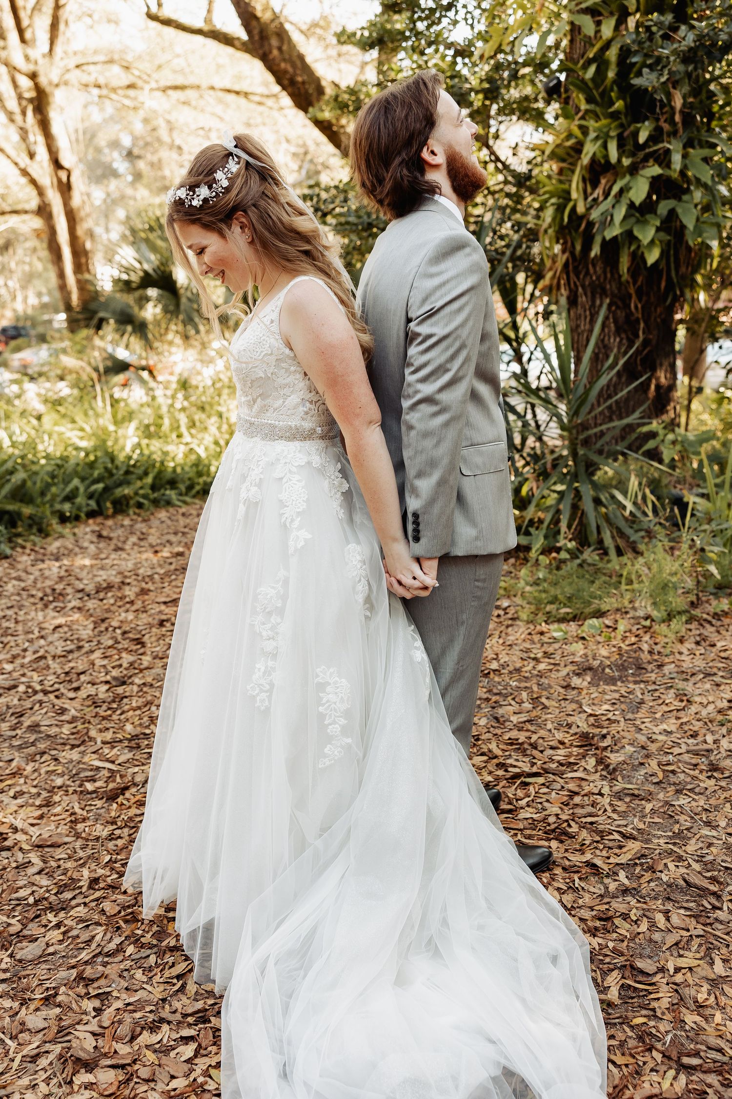 A couple in wedding attire stands back to back holding hands in a garden setting.