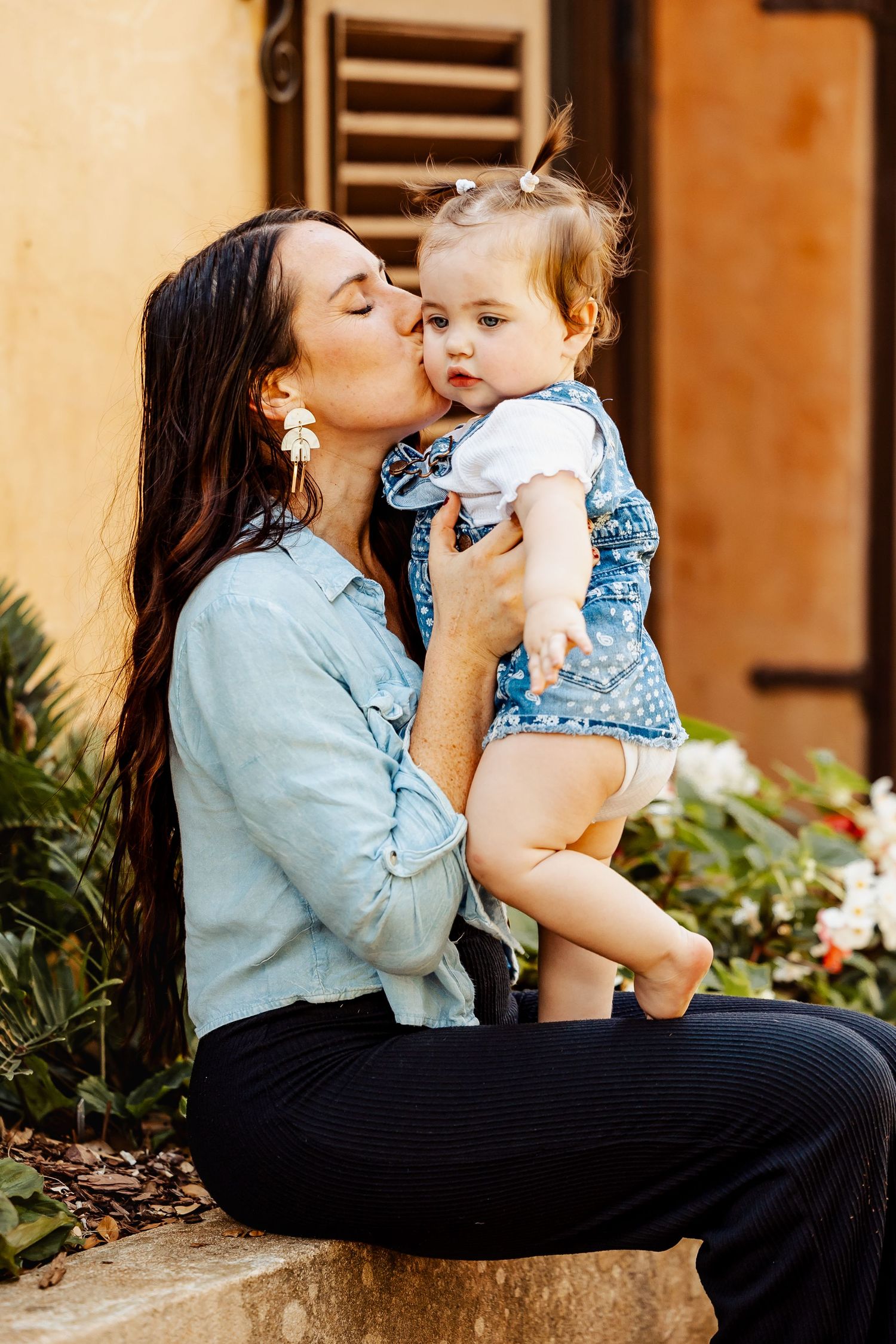 Loving mother kissing her baby girl on the cheek while sitting outdoors near colorful flowers.
