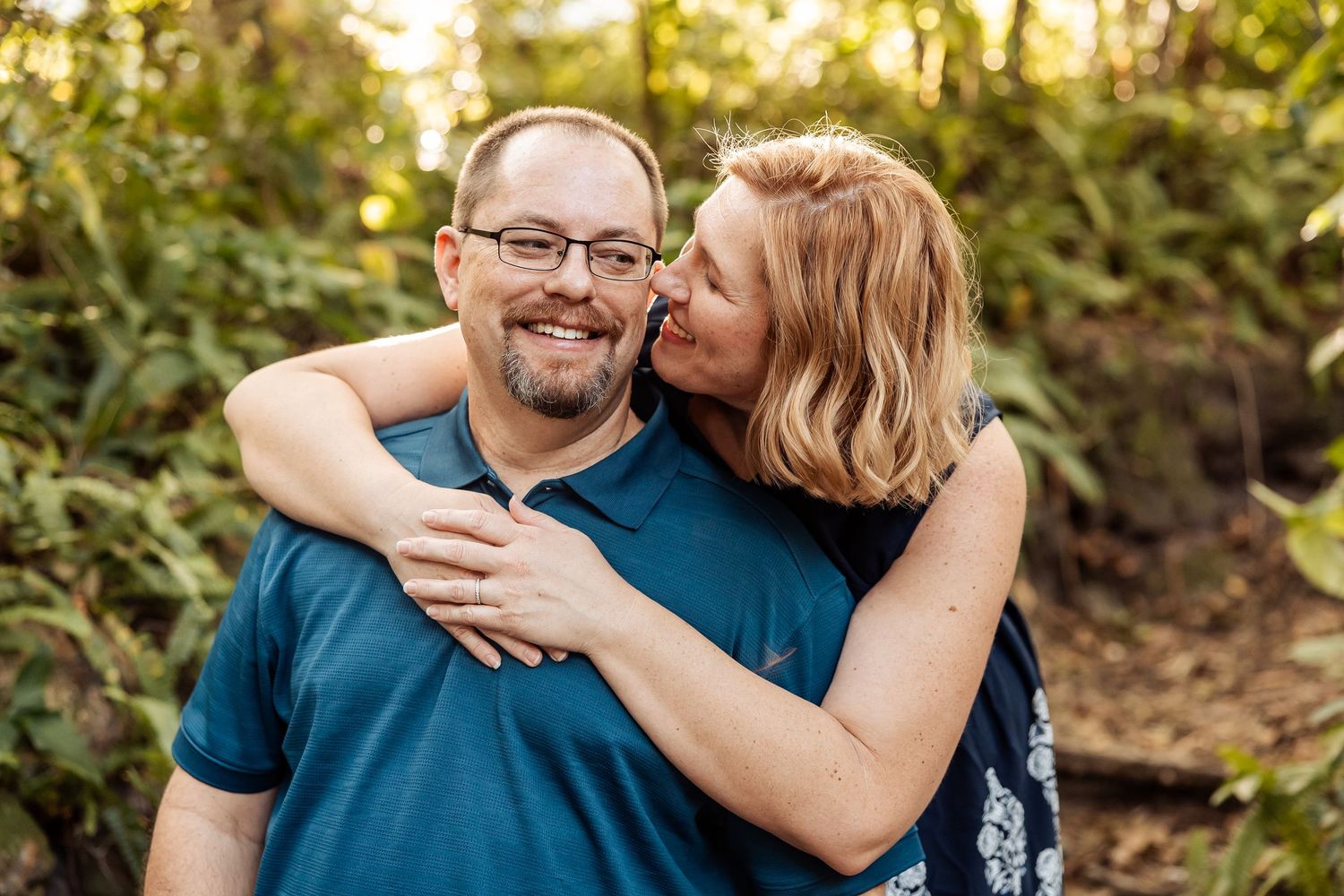 Couple embracing outdoors, woman kissing man's cheek while he smiles, surrounded by lush green foliage.