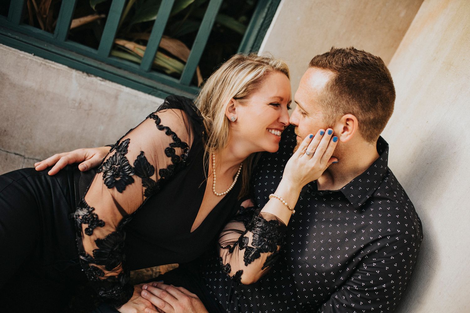 Romantic couple in black outfits sharing an intimate moment while seated against a wall outdoors.
