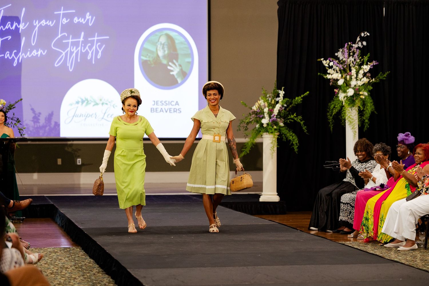 Two women in vintage green dresses walk runway holding hands at fashion show.
