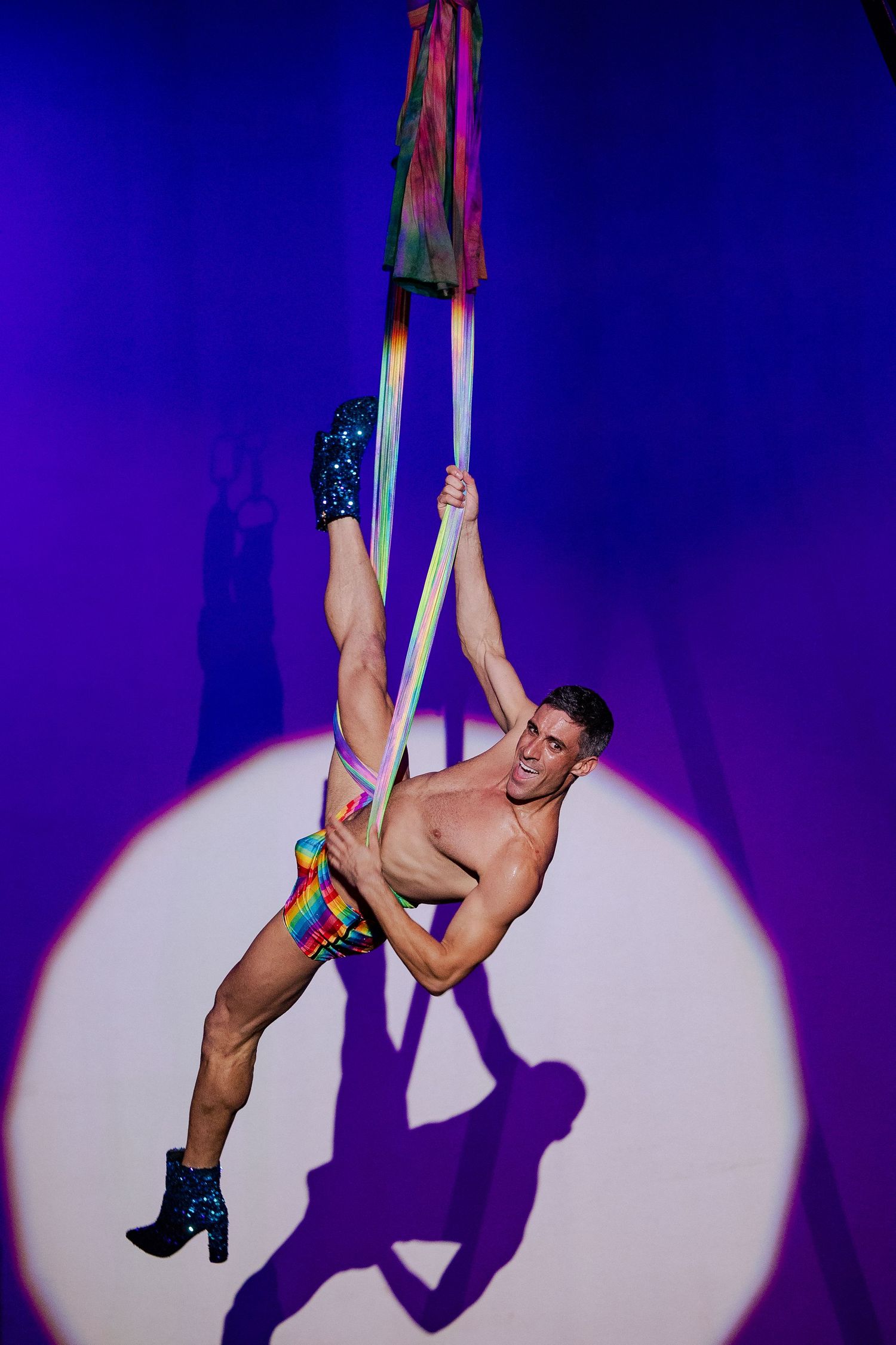 Athletic shirtless male aerialist performing on aerial silks in rainbow shorts and glitter boots.