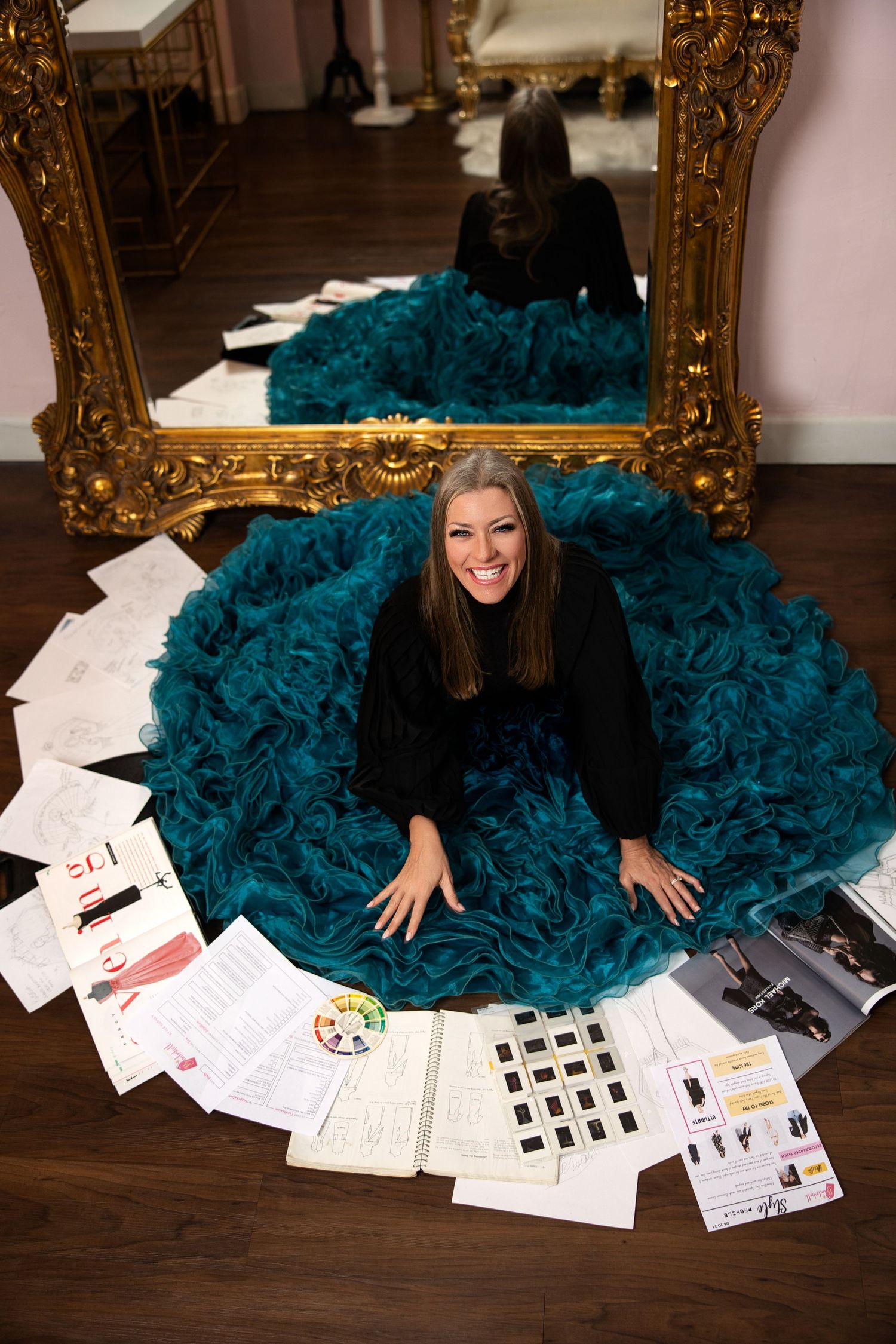 Woman in black top sitting in a teal ruffled skirt surrounded by design sketches near a gold mirror.