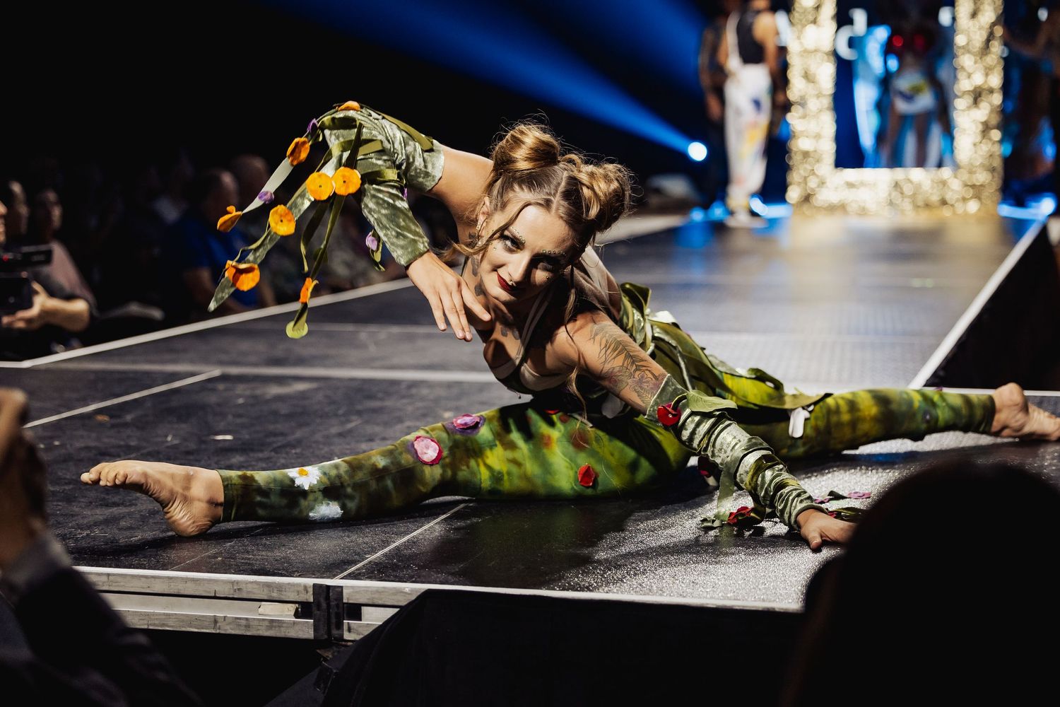 Flexible performer in camouflage outfit doing a floor split on a runway stage.