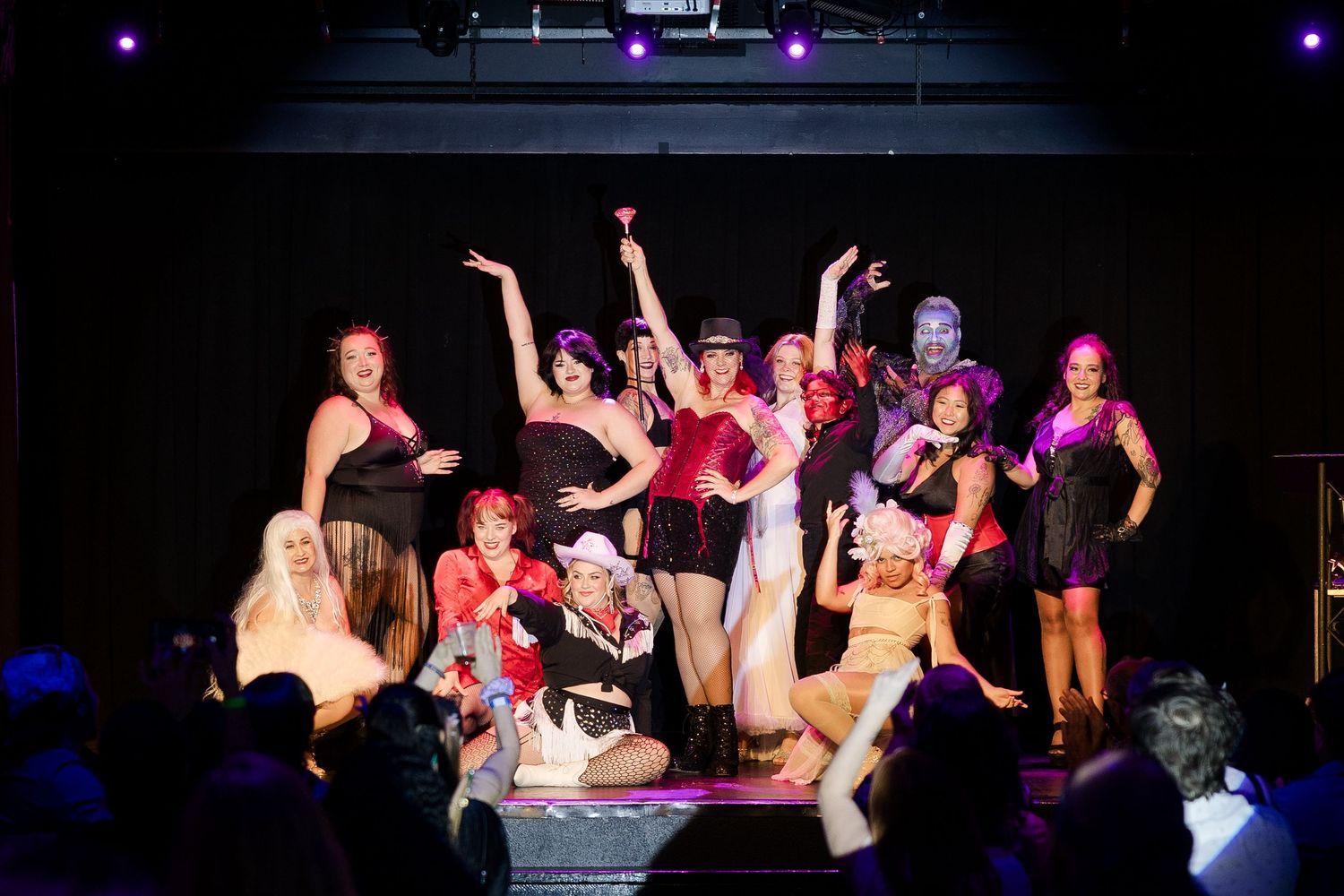 Large group of burlesque performers posing together on dark stage after show finale.