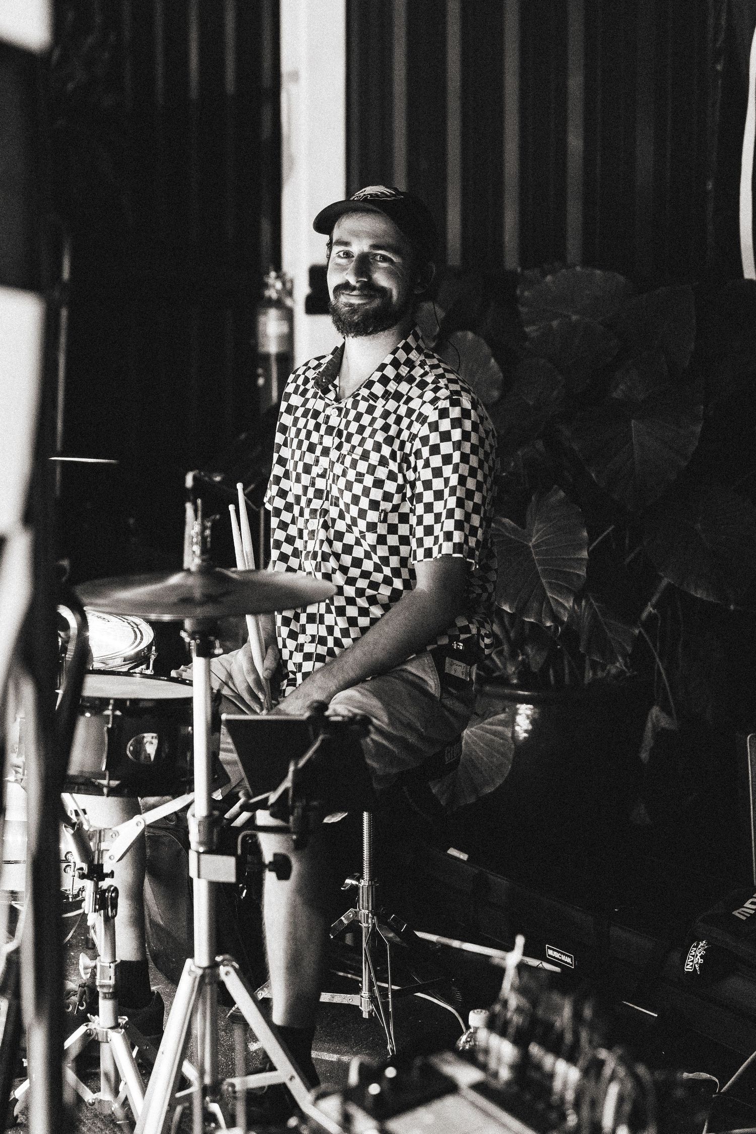 A smiling bearded drummer in a checkered shirt sits behind his drum kit in a black and white photo.