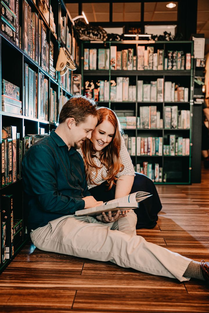 Couple sitting on wooden floor in bookstore, smiling and reading together surrounded by colorful shelves of books.