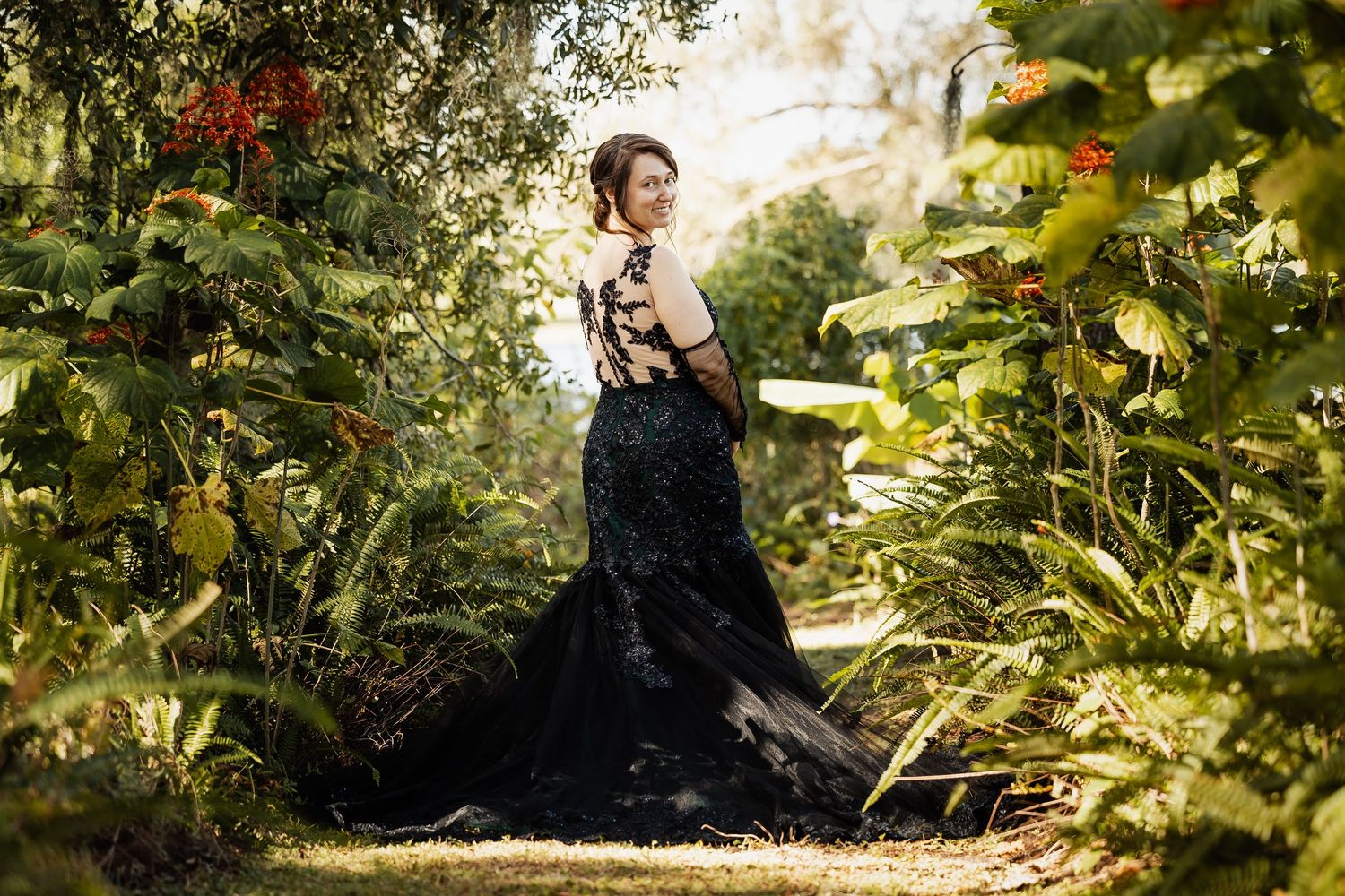 Woman in stunning black lace gown poses elegantly among lush tropical garden greenery.