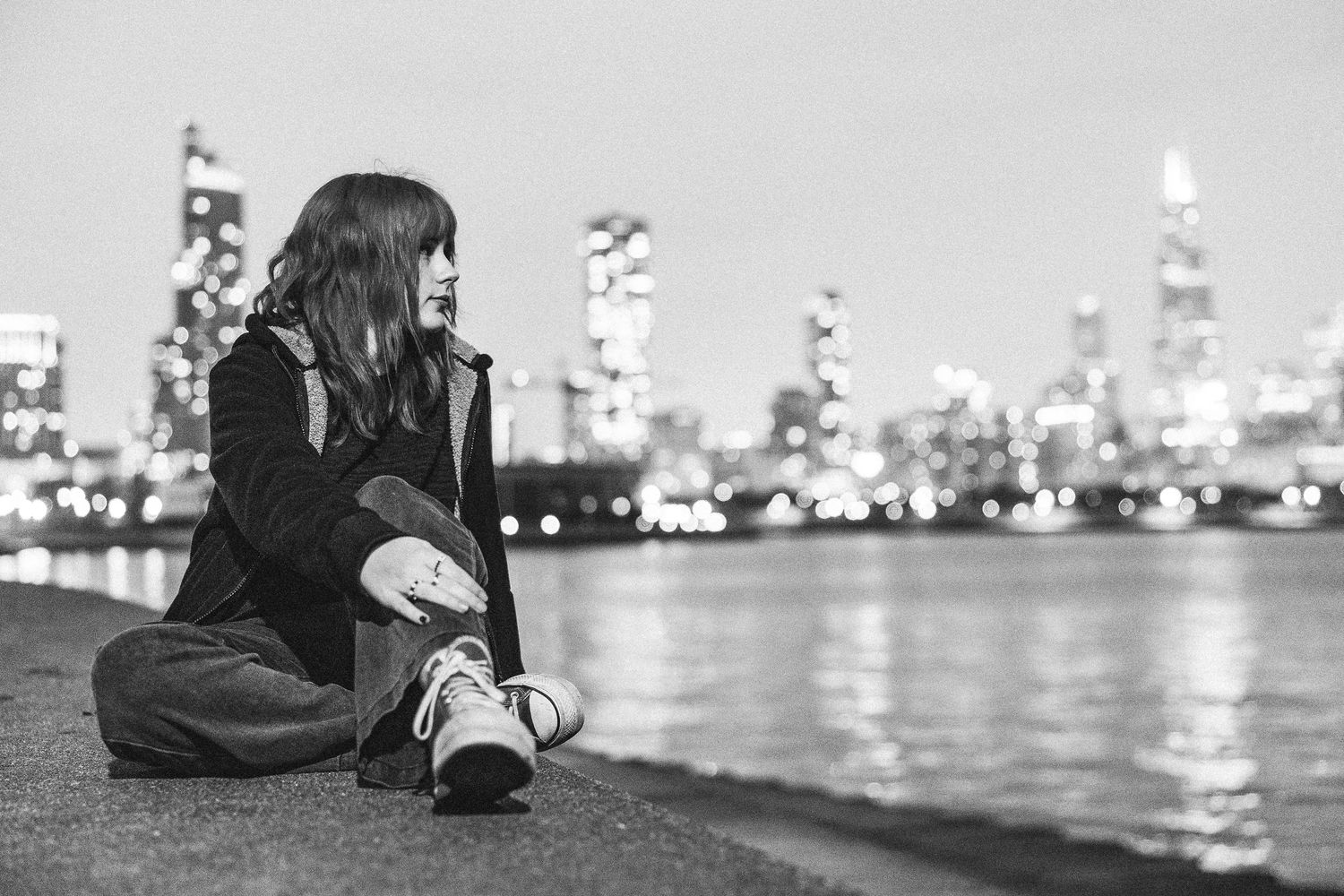 Black and white photo of a young woman sitting by a city waterfront at night with bokeh lights in the background.