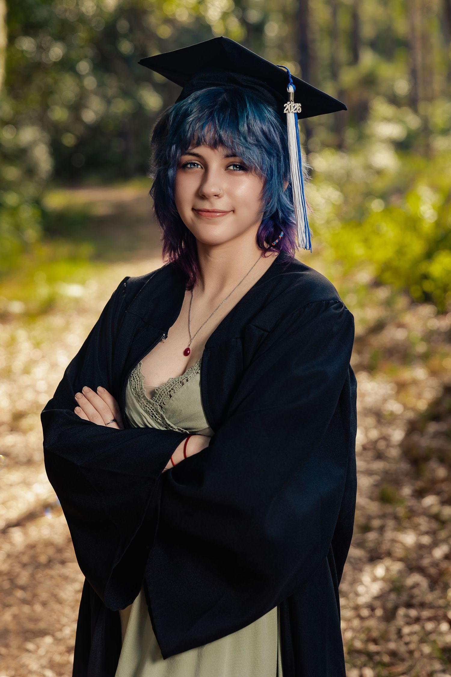 Young woman with blue hair in graduation cap and gown, arms crossed, smiling outdoors in a sunlit forest setting.