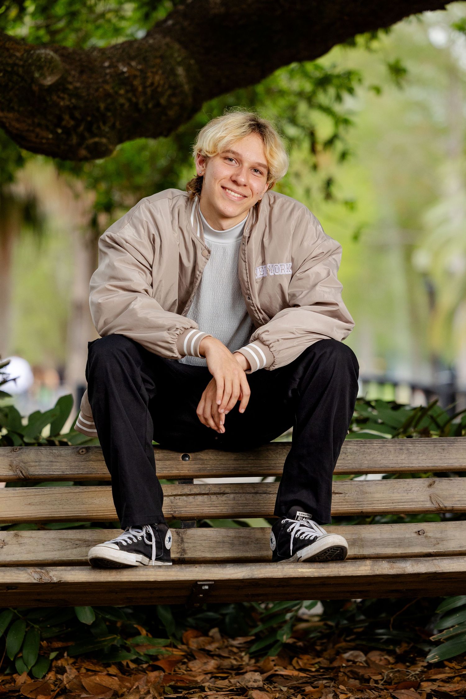 Smiling teen boy in beige bomber jacket sitting on a wooden bench under a tree in a park.