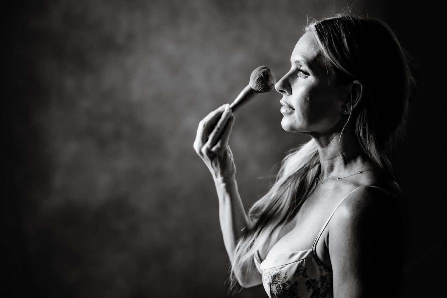 Black and white portrait of a woman applying makeup with a brush, captured in dramatic studio lighting.