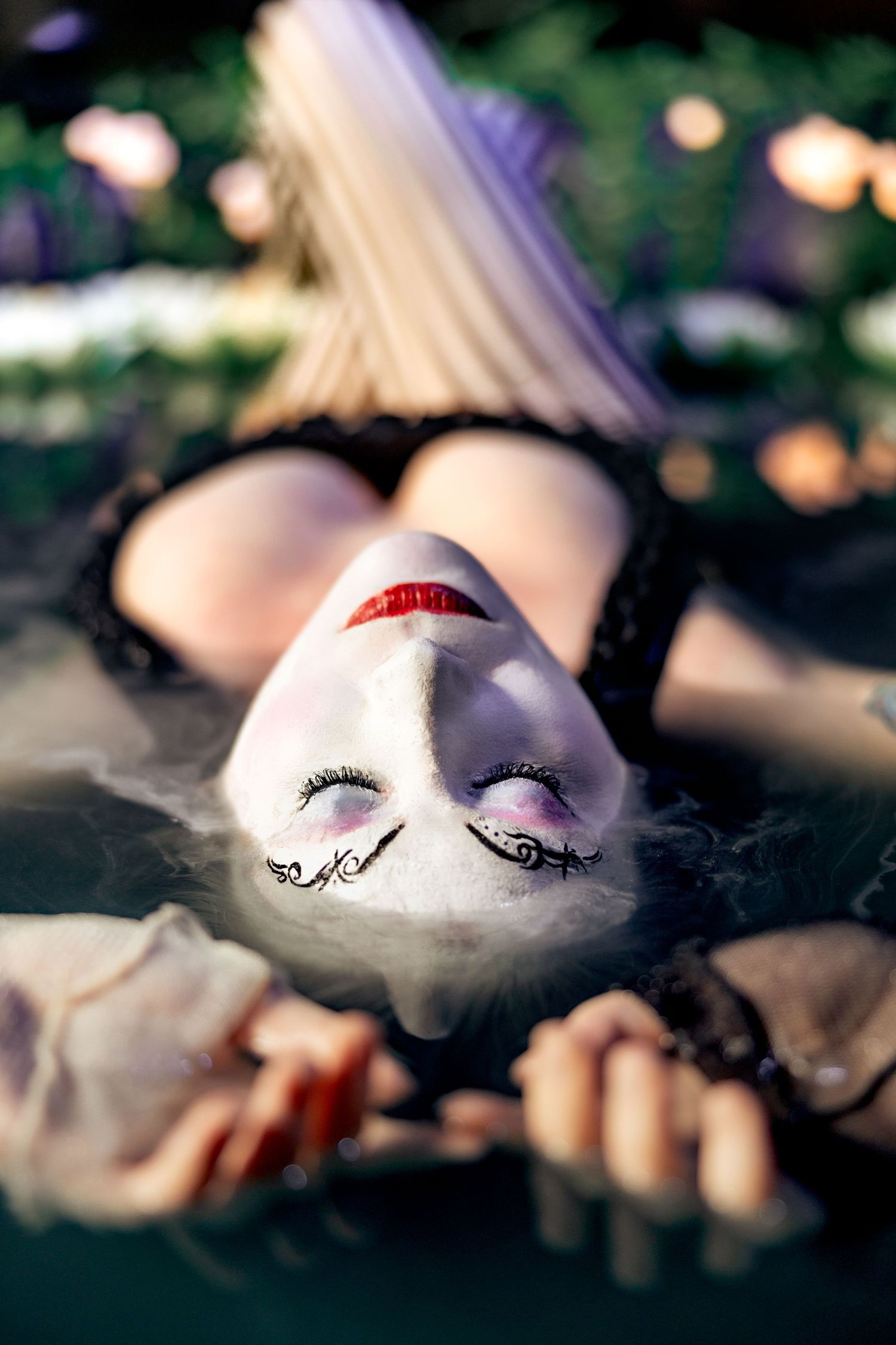 A woman with dramatic white face paint and gothic makeup lies on the ground surrounded by greenery.