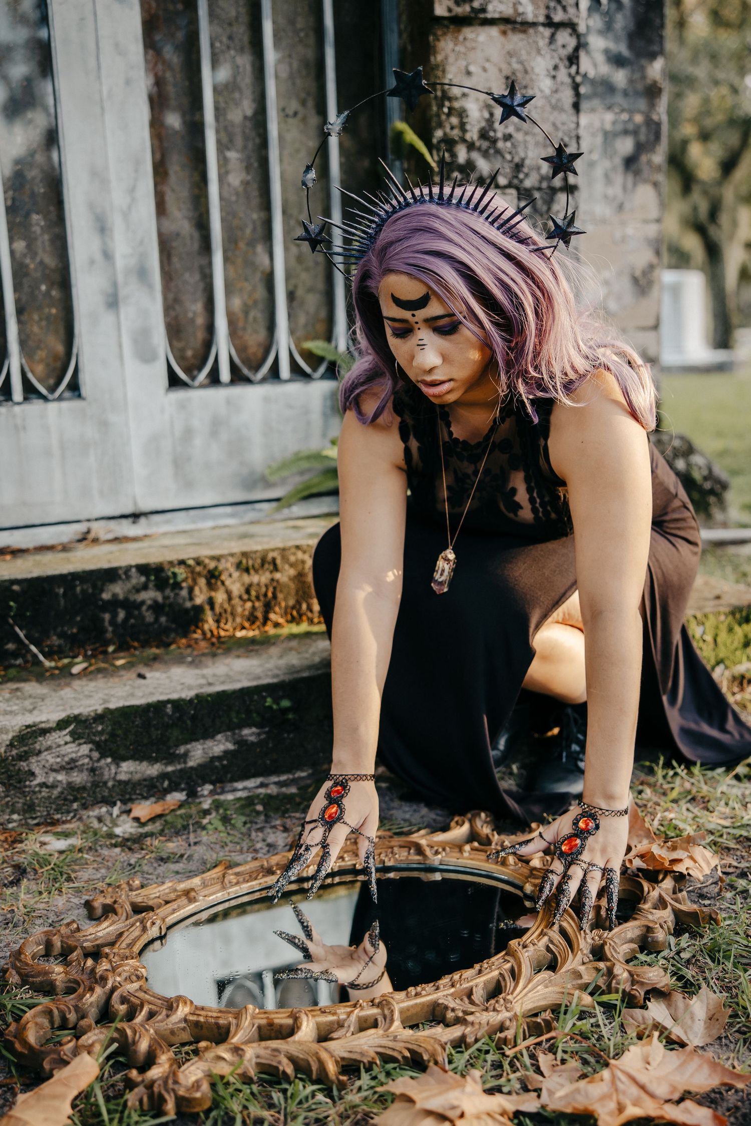 A woman with purple hair and star crown crouches over an ornate mirror on the ground outdoors.
