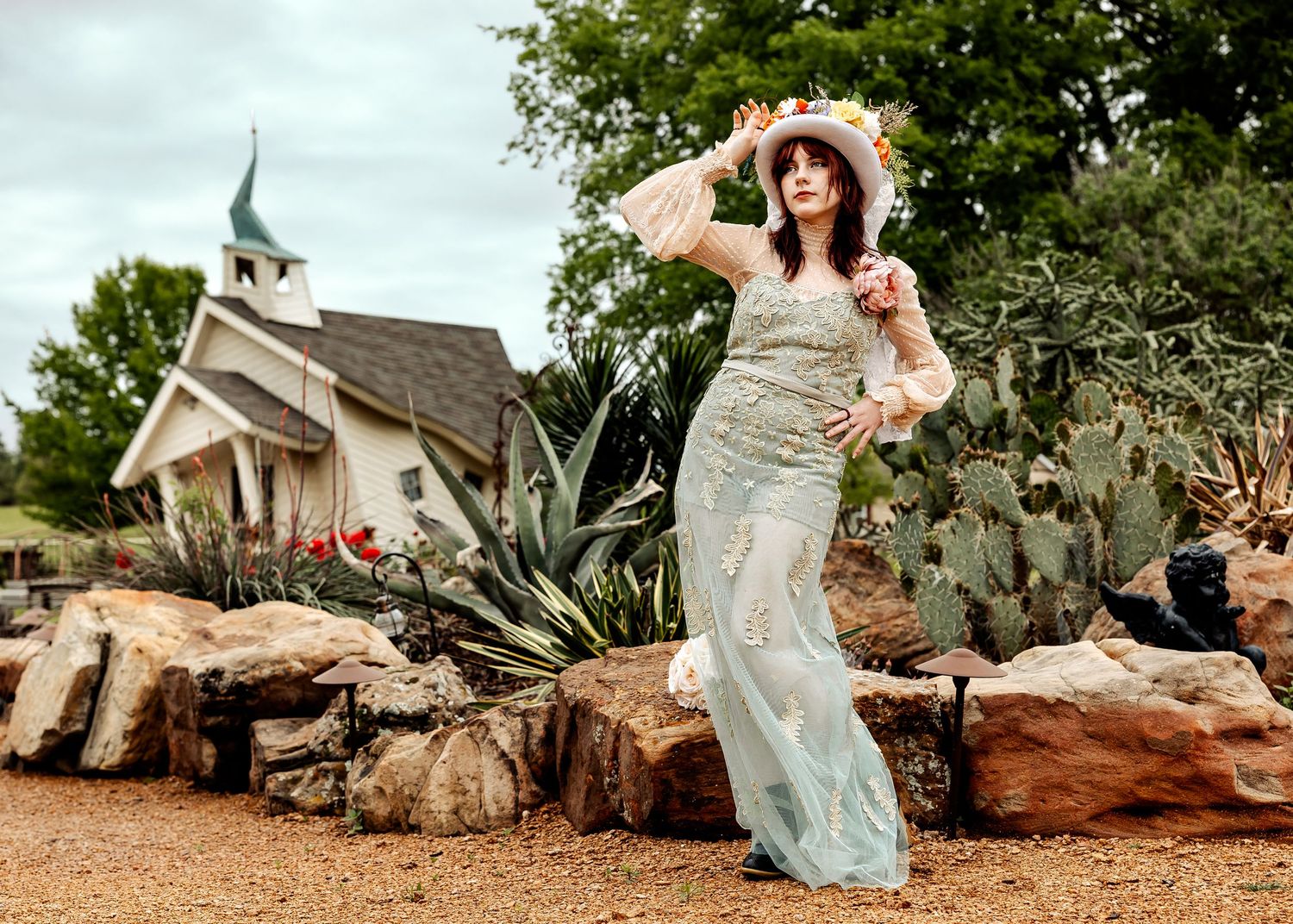 Woman in mint lace gown and floral hat poses among rocks and cacti near a white chapel.