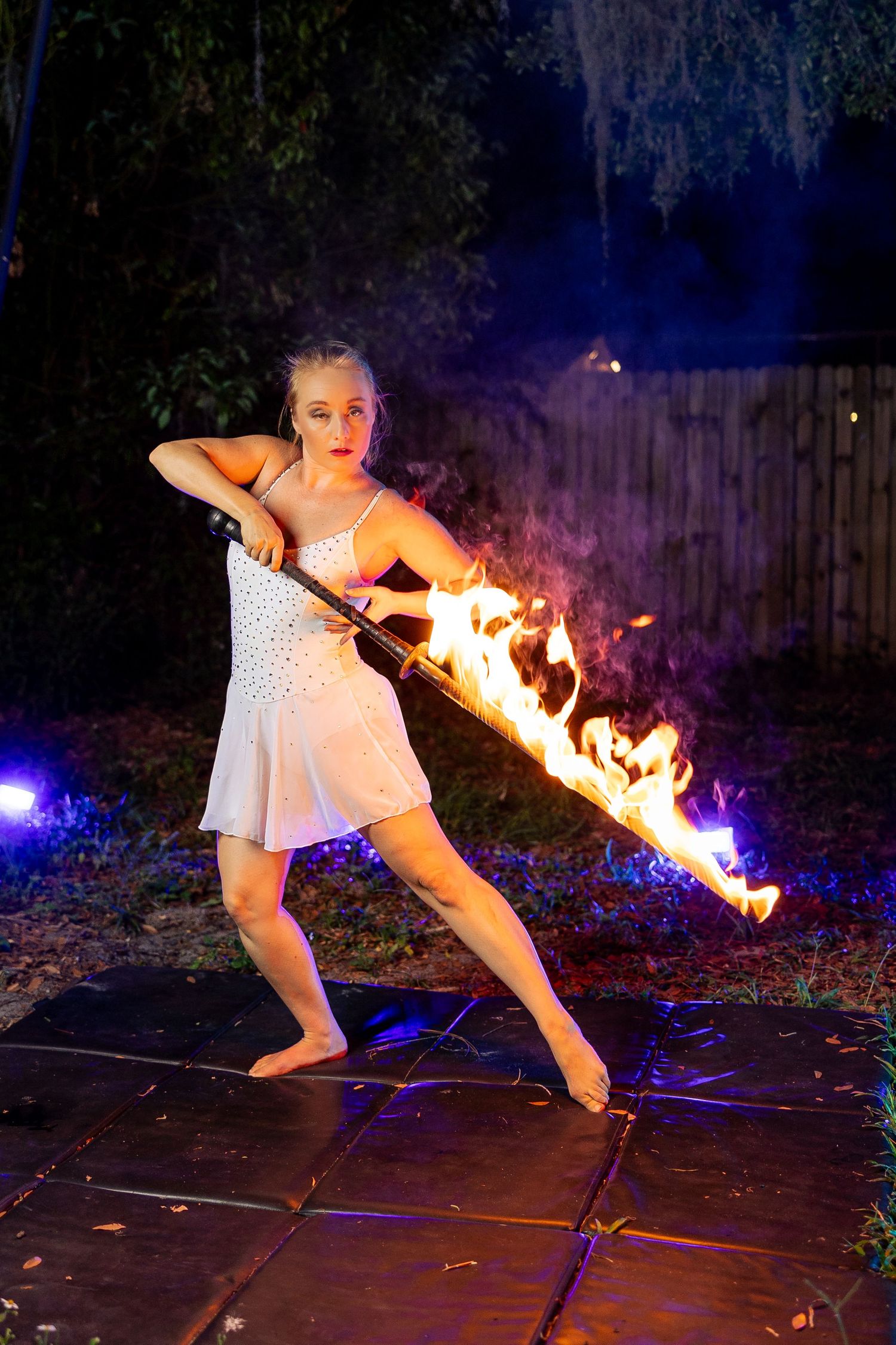 Female fire performer in white dress wielding a flaming staff outdoors at night with blue lighting.