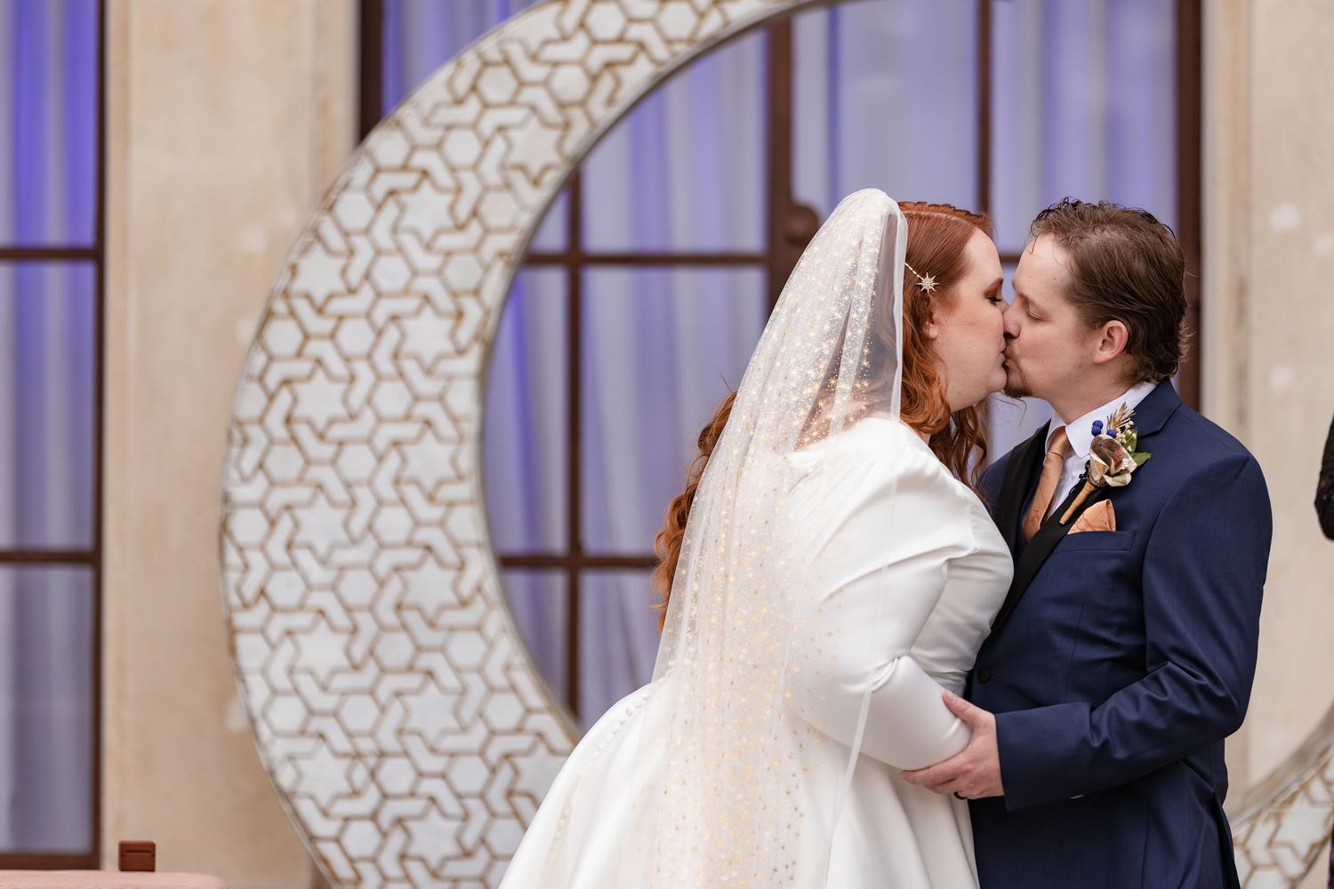 Bride and groom share first kiss in front of decorative crescent moon ceremony backdrop.