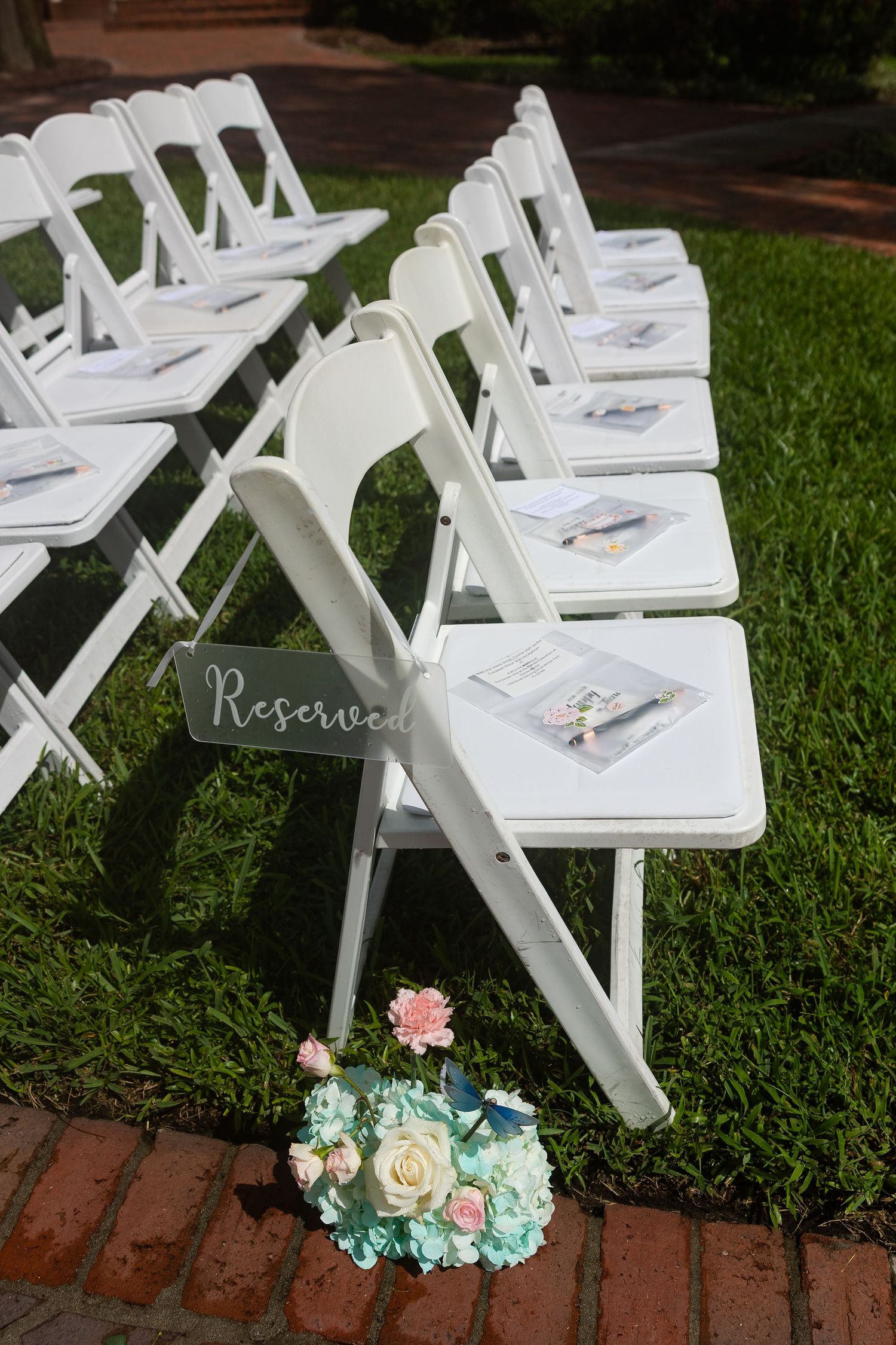 White folding chairs with programs set up outdoors for a wedding ceremony with a Reserved sign and flowers.