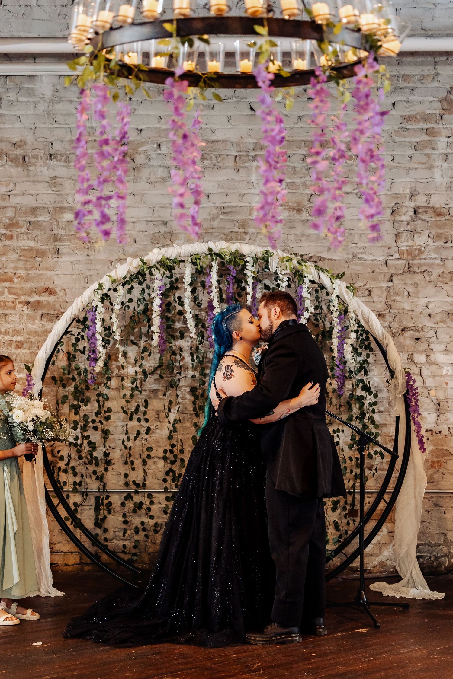 Bride in black sequin gown and groom share first kiss before circular floral arch with wisteria.