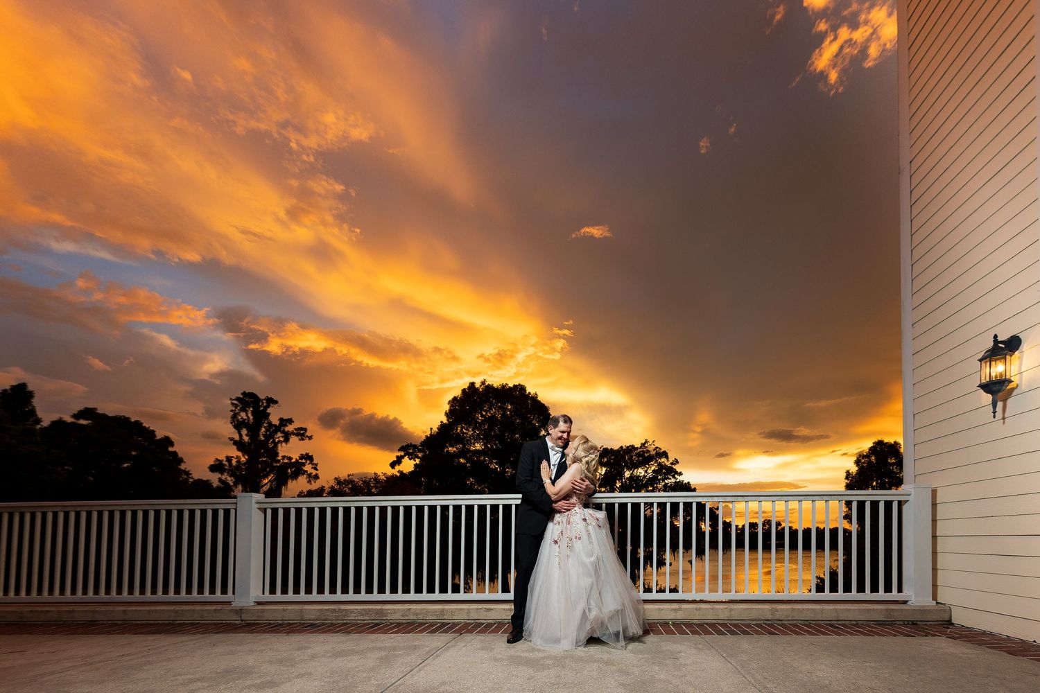 Bride and groom pose on a balcony against a dramatic golden sunset sky on their wedding day.