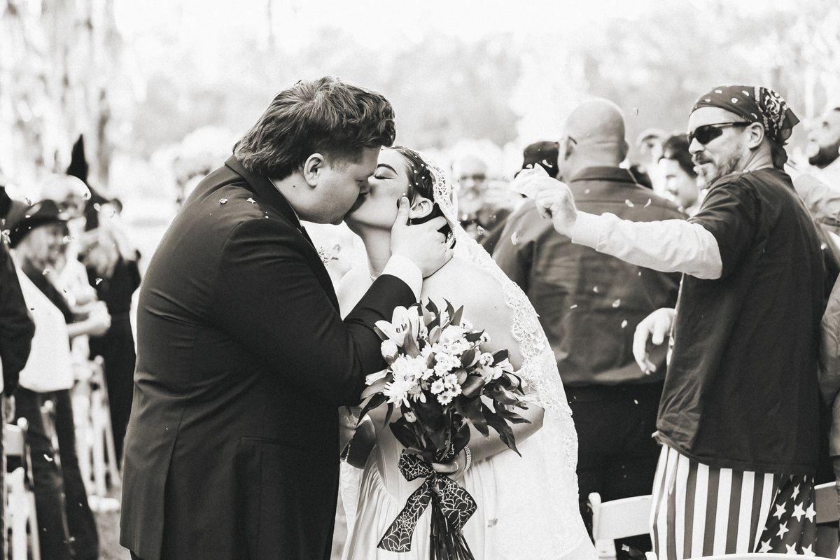 Bride and groom share first kiss at altar as guests celebrate with confetti, black and white.
