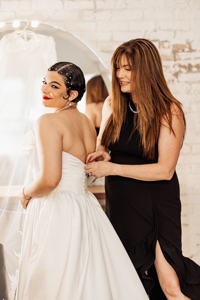 Bridesmaid in black dress helping bride button up white strapless wedding gown in bright studio.